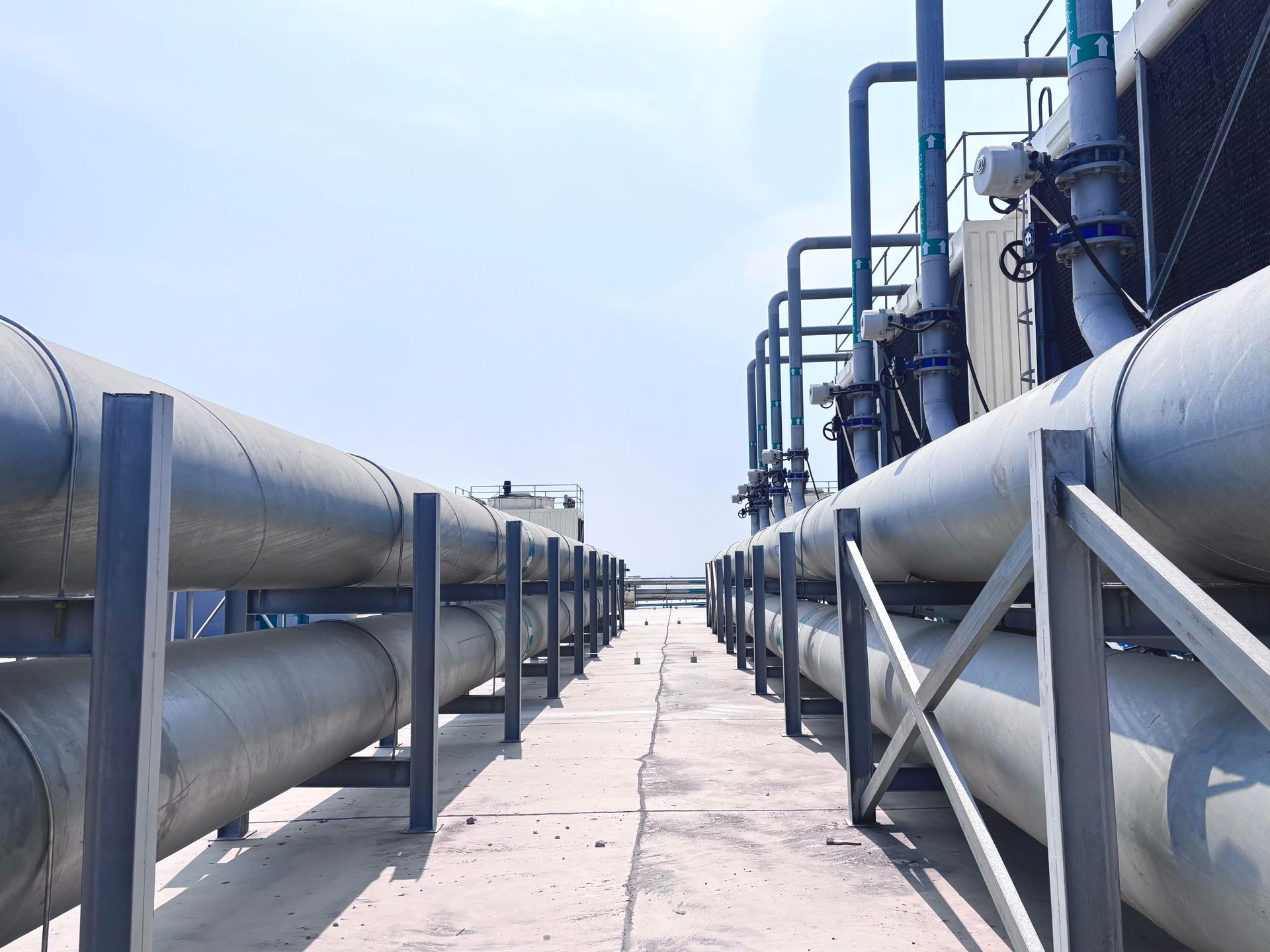 Industrial walkway between large silver pipes and steel supports under a blue sky