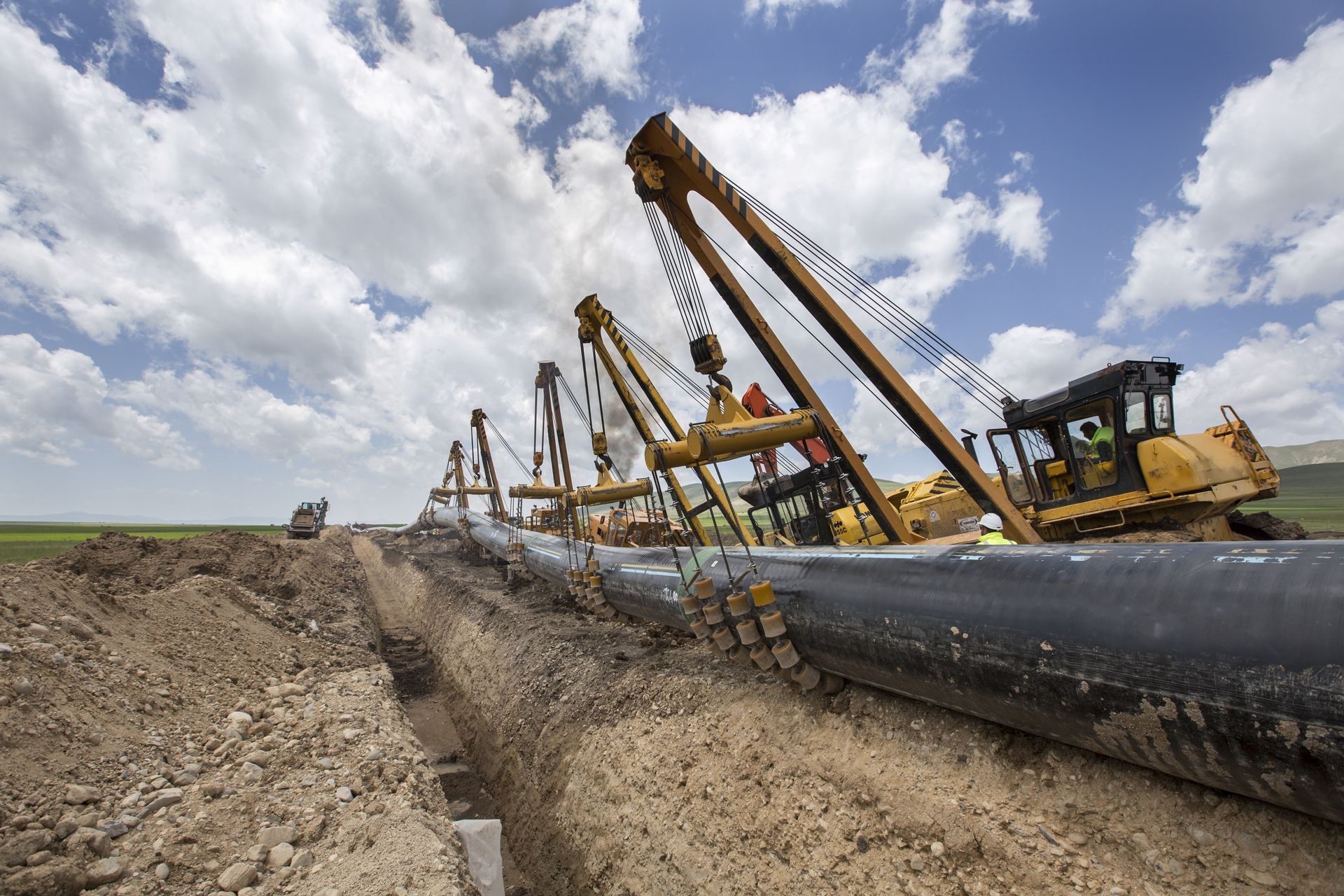 Pipeline construction machine laying large black pipe along a dirt trench under a cloudy sky