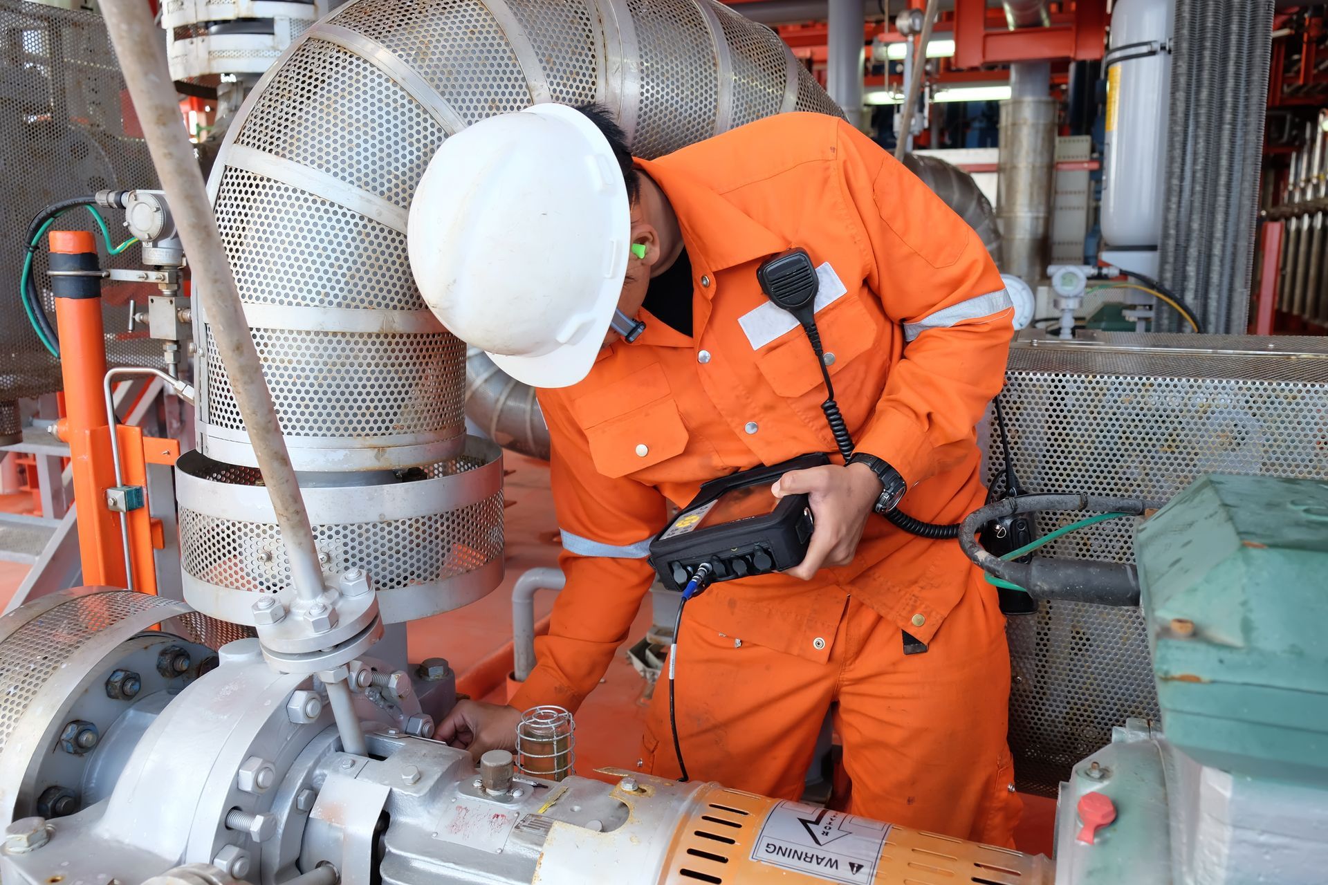 Worker in orange coveralls and hard hat inspecting industrial piping in a factory setting