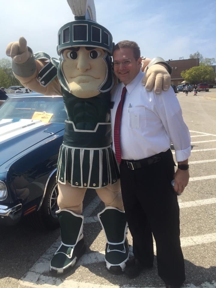 Man poses with Sparty mascot, pointing. Outdoor setting with a vintage car visible.