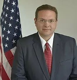 Man in suit and tie stands near US flag.