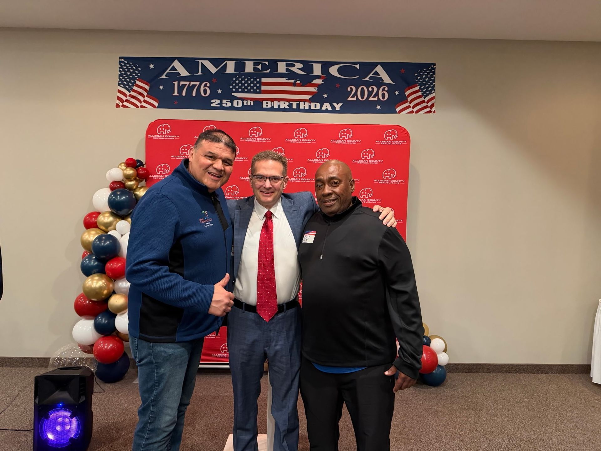 Three men posing at a patriotic event with red, white, and blue decorations and an America 2026 banner