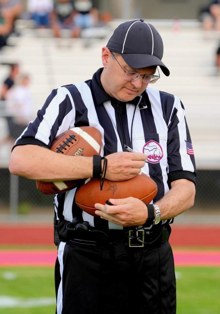 Football referee holding two footballs on a football field. He wears a black and white striped shirt and a hat.
