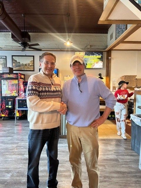 Two men shaking hands inside a casual restaurant. One wears a patterned sweater, the other a polo shirt. A server walks by.