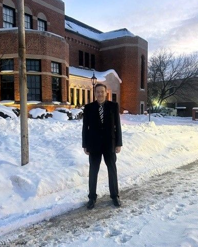 Man in black suit stands on snow-covered sidewalk in front of a brick building.