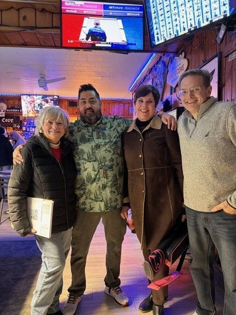 Four people pose together inside a bar with a TV screen above them.