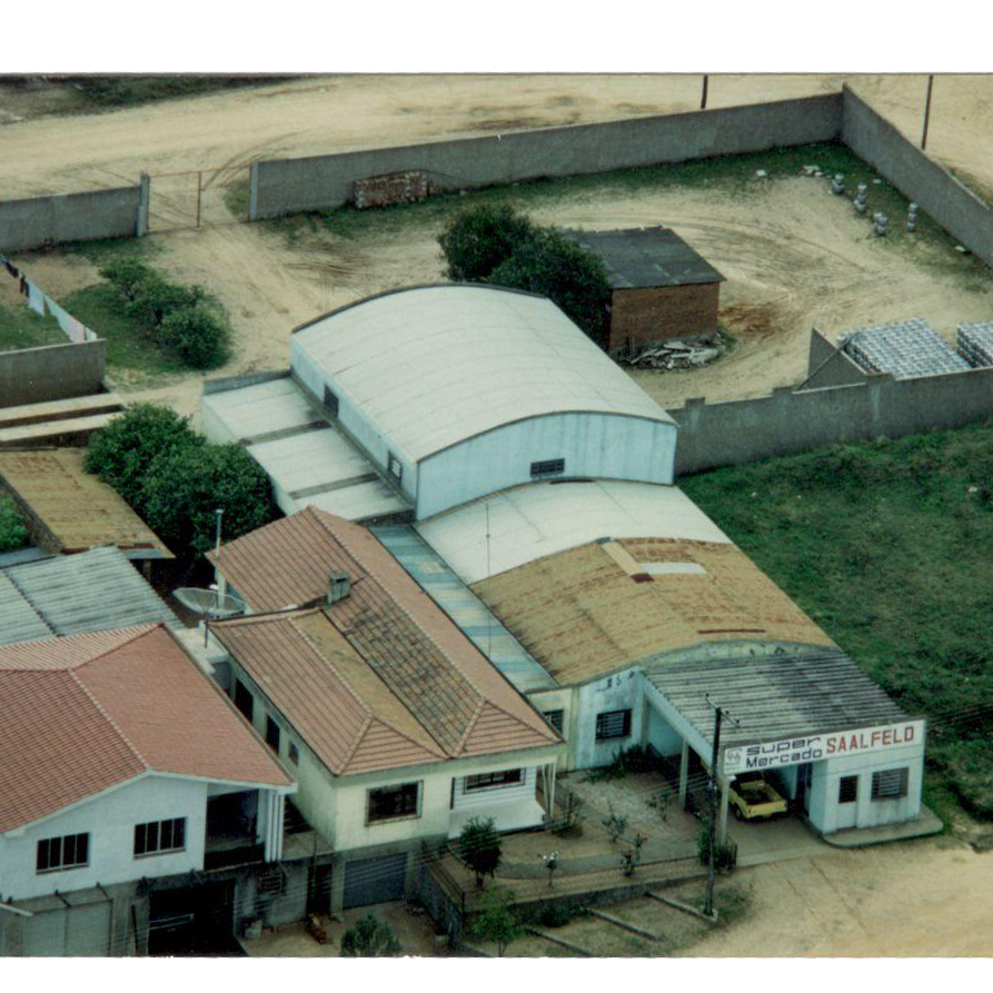 Vista aérea de um pequeno complexo com casas de telha, um grande armazém com telhado curvo e um terreno de terra cercado.