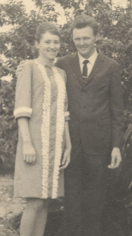 Um casal sorridente posa lado a lado ao ar livre em um retrato antigo em preto e branco.