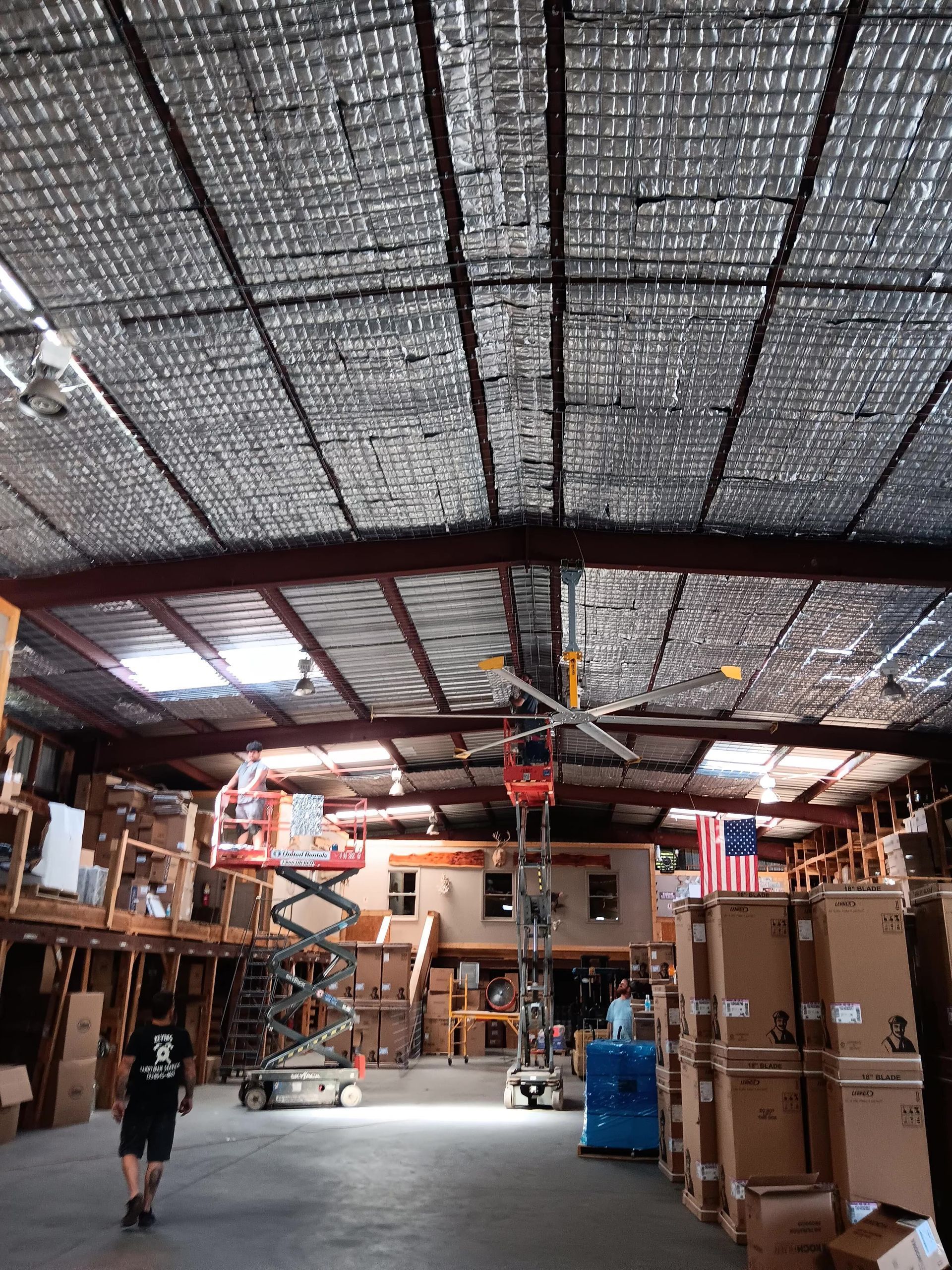 Warehouse interior: workers on lifts installing silver insulation on the metal ceiling, surrounded by stacked boxes.