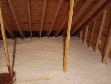 Attic with white insulation between wooden rafters, under a plywood roof.