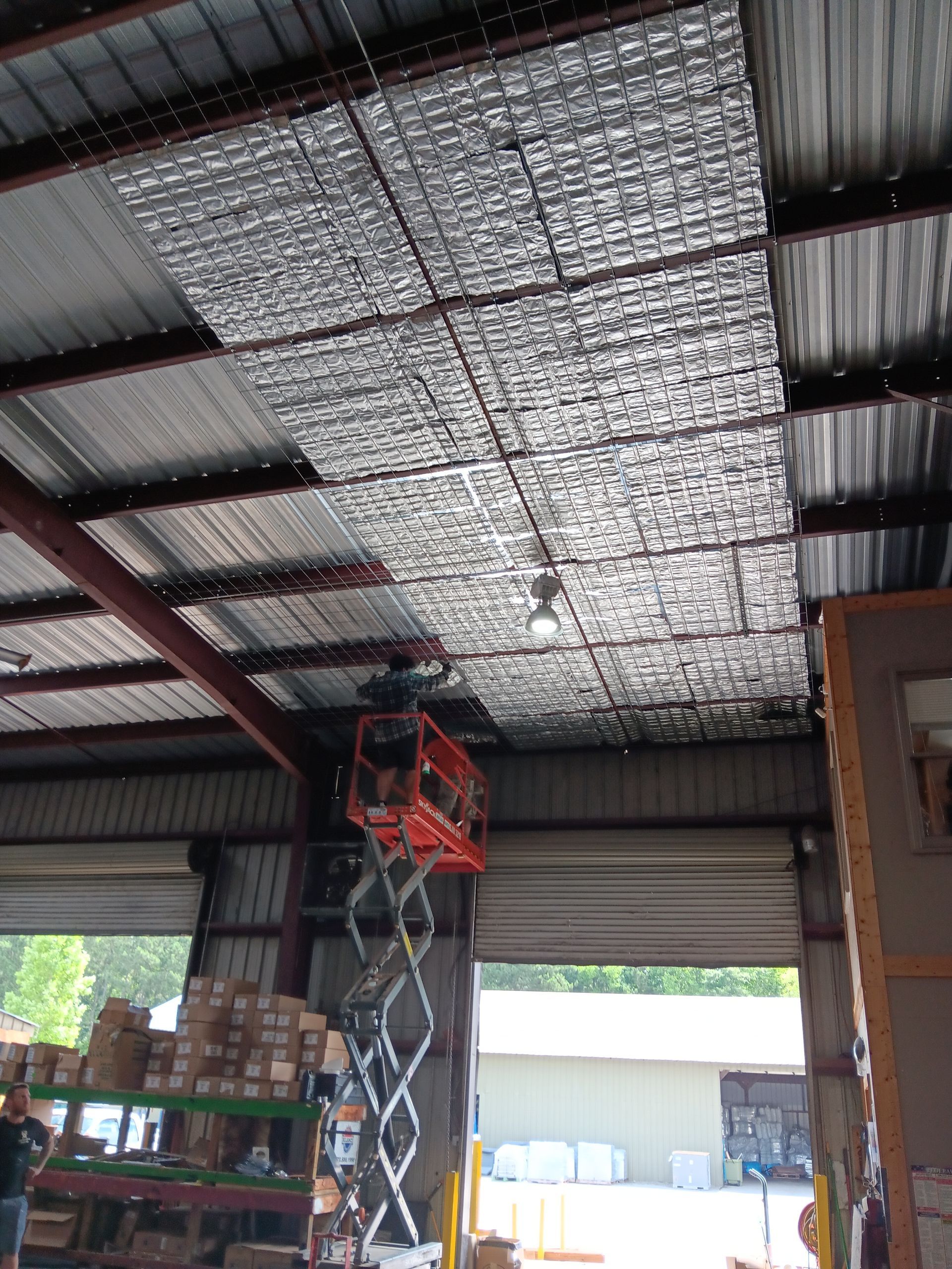 Warehouse interior with a scissor lift under a corrugated metal ceiling, silver insulation panels installed.