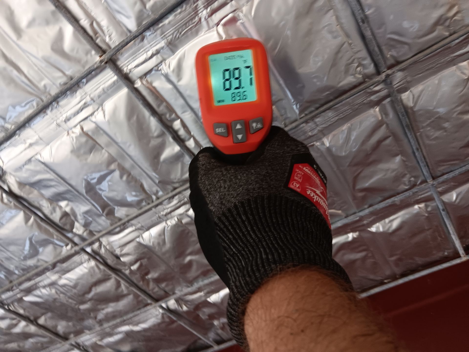 Hand in work glove holding thermometer measuring ceiling temperature, reading 89.7°F.