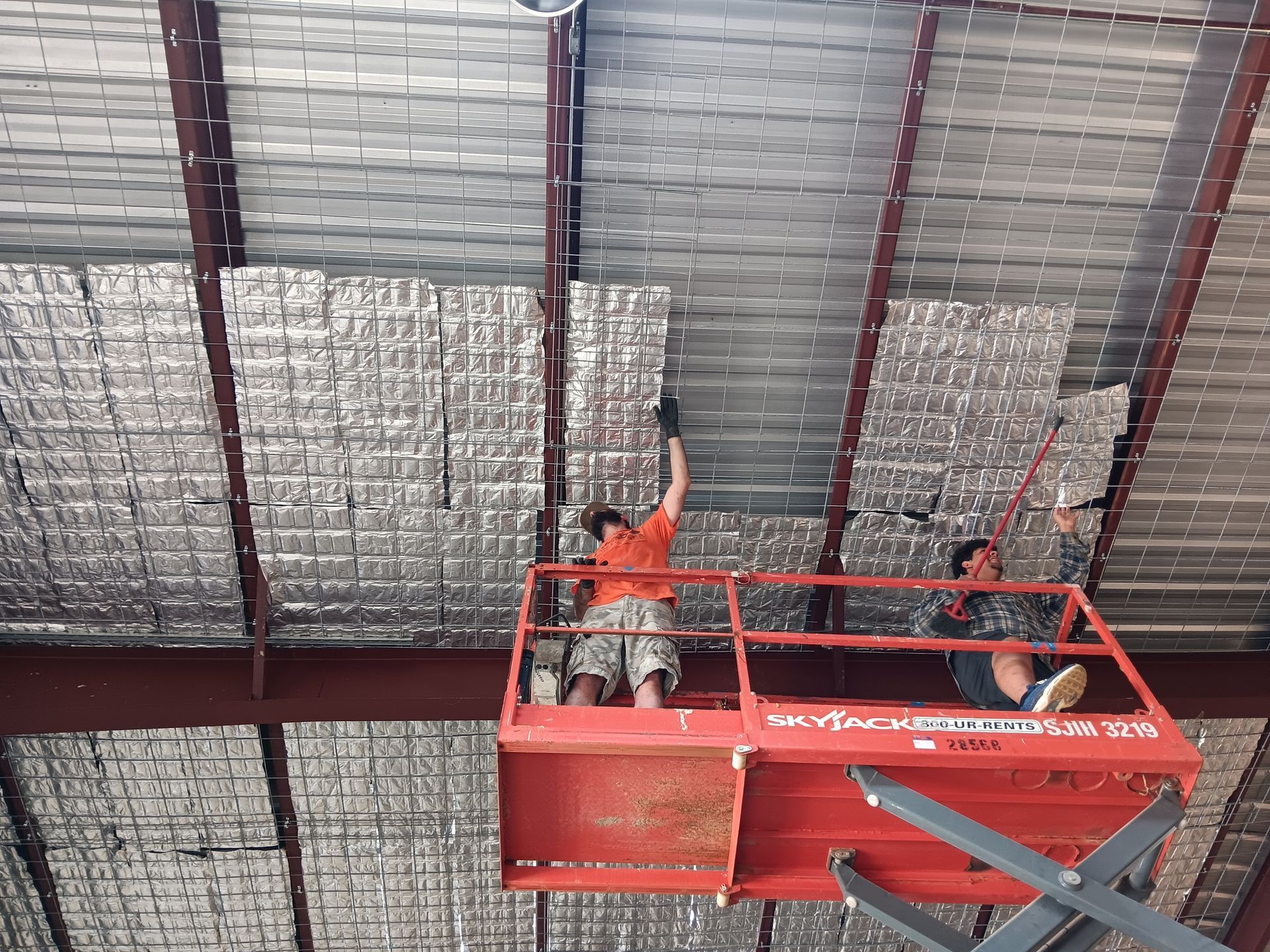 Two workers installing insulation on a metal ceiling from an elevated platform.