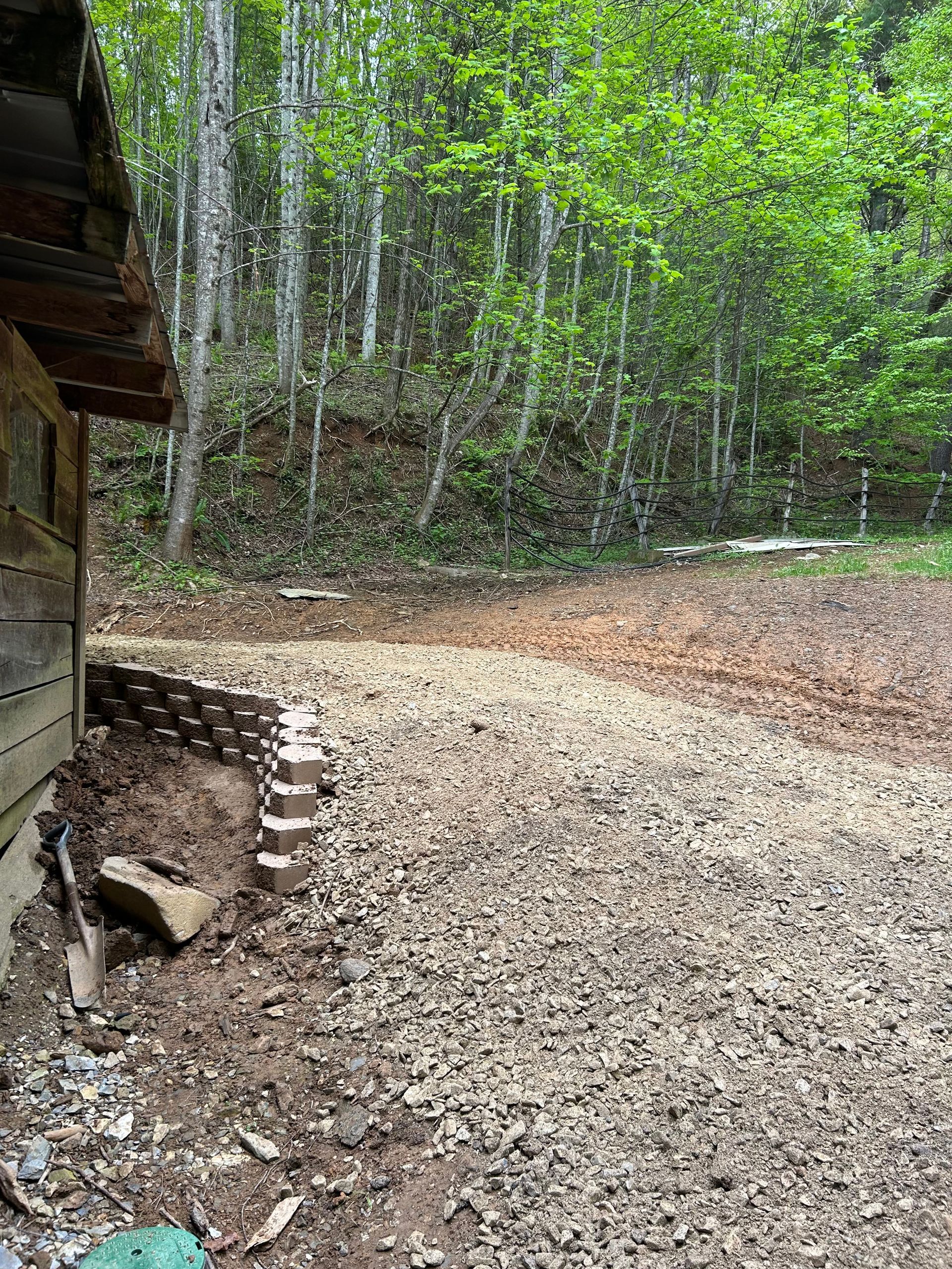 Wooded outdoor area beside a rustic cabin, with gravel ground and green trees in the background.