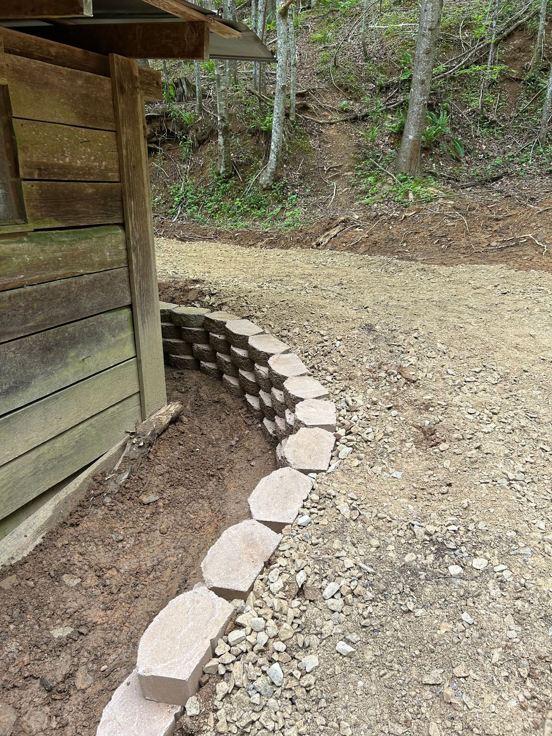 Curved stone edging along a gravel path beside a wooden structure in a wooded area