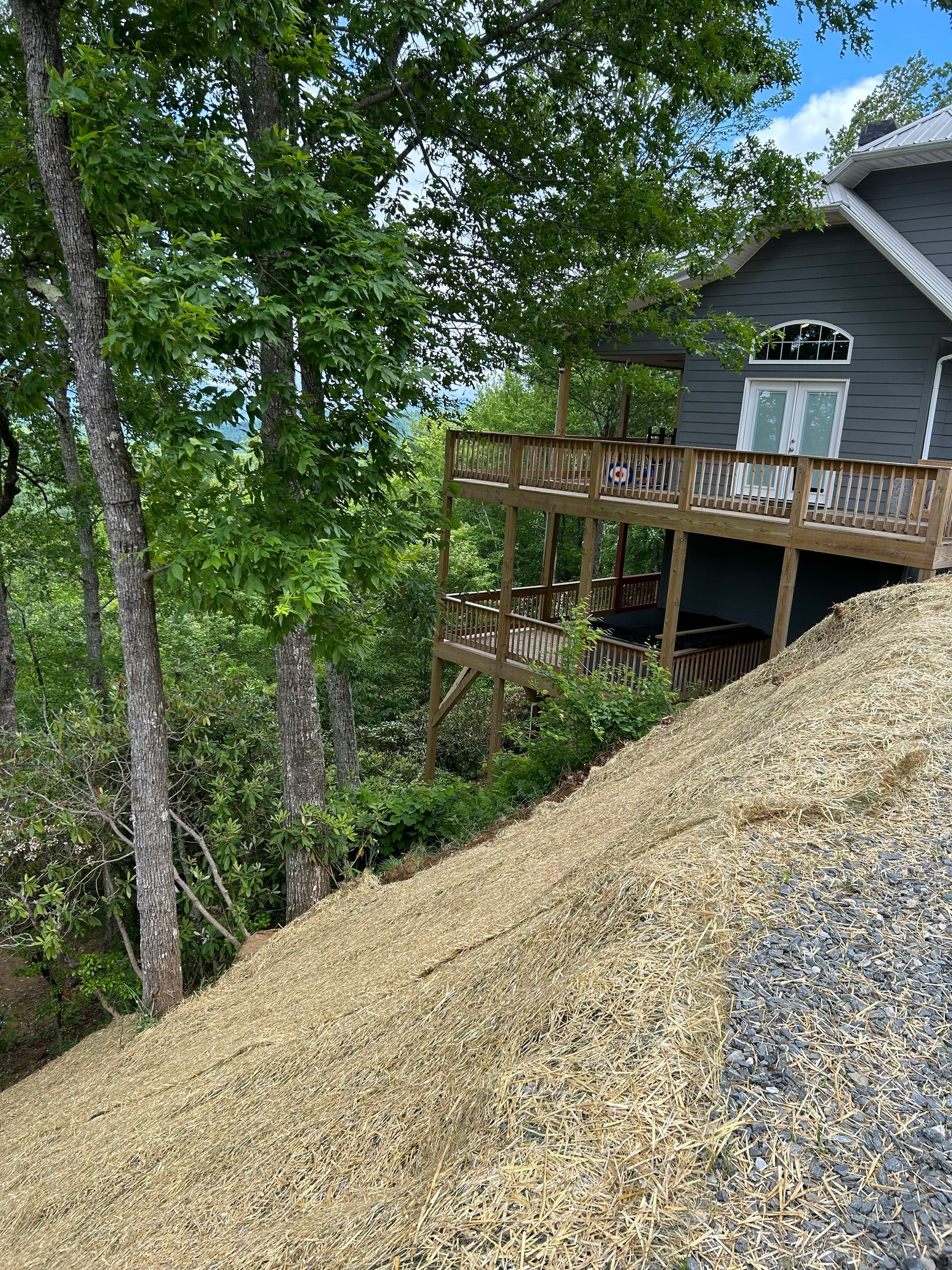 Steep gravel pile beside a house deck, with trees and a wooded yard in the background