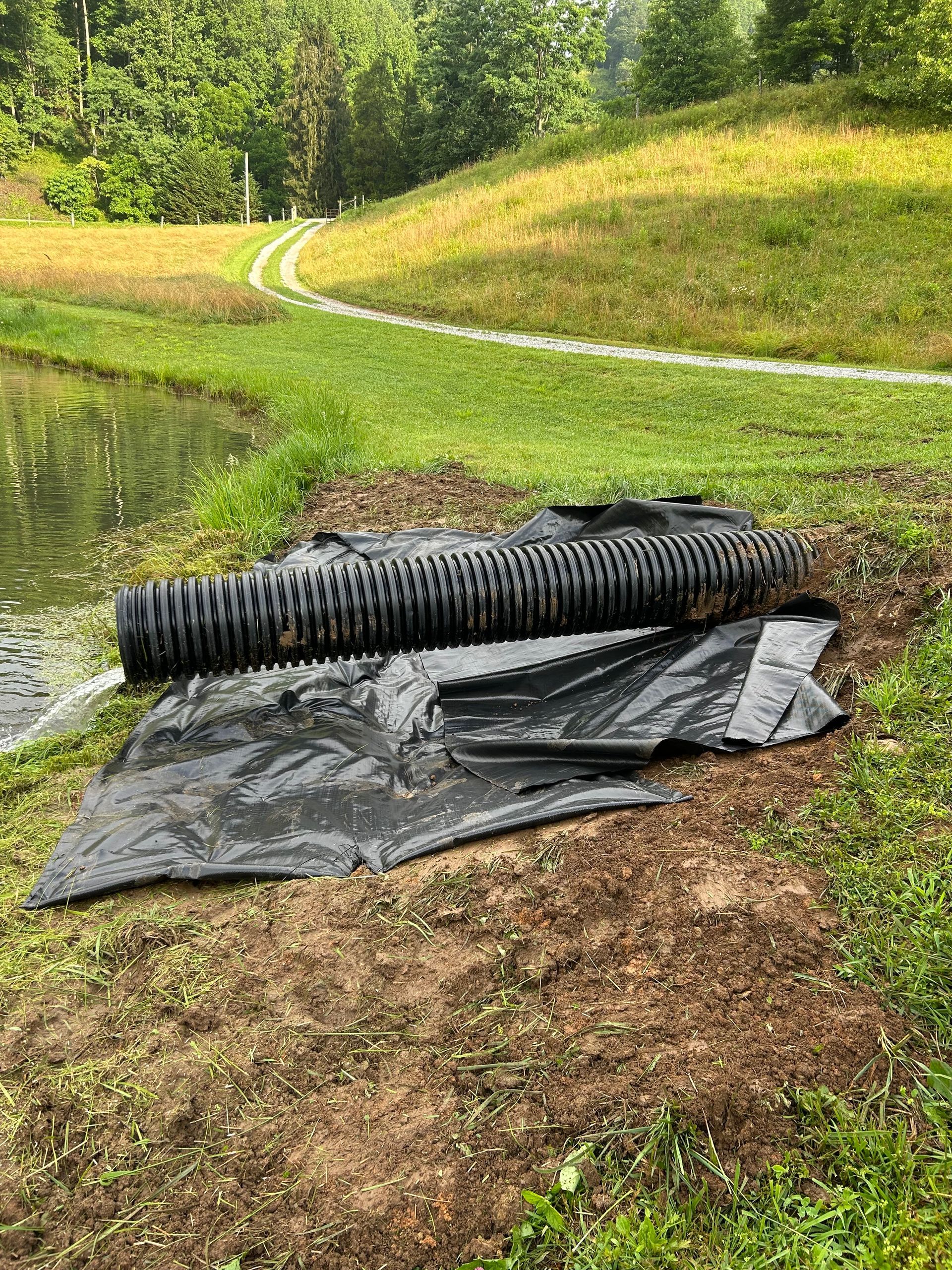 Black drainage pipe and tarp beside a muddy pond in a grassy, wooded area.