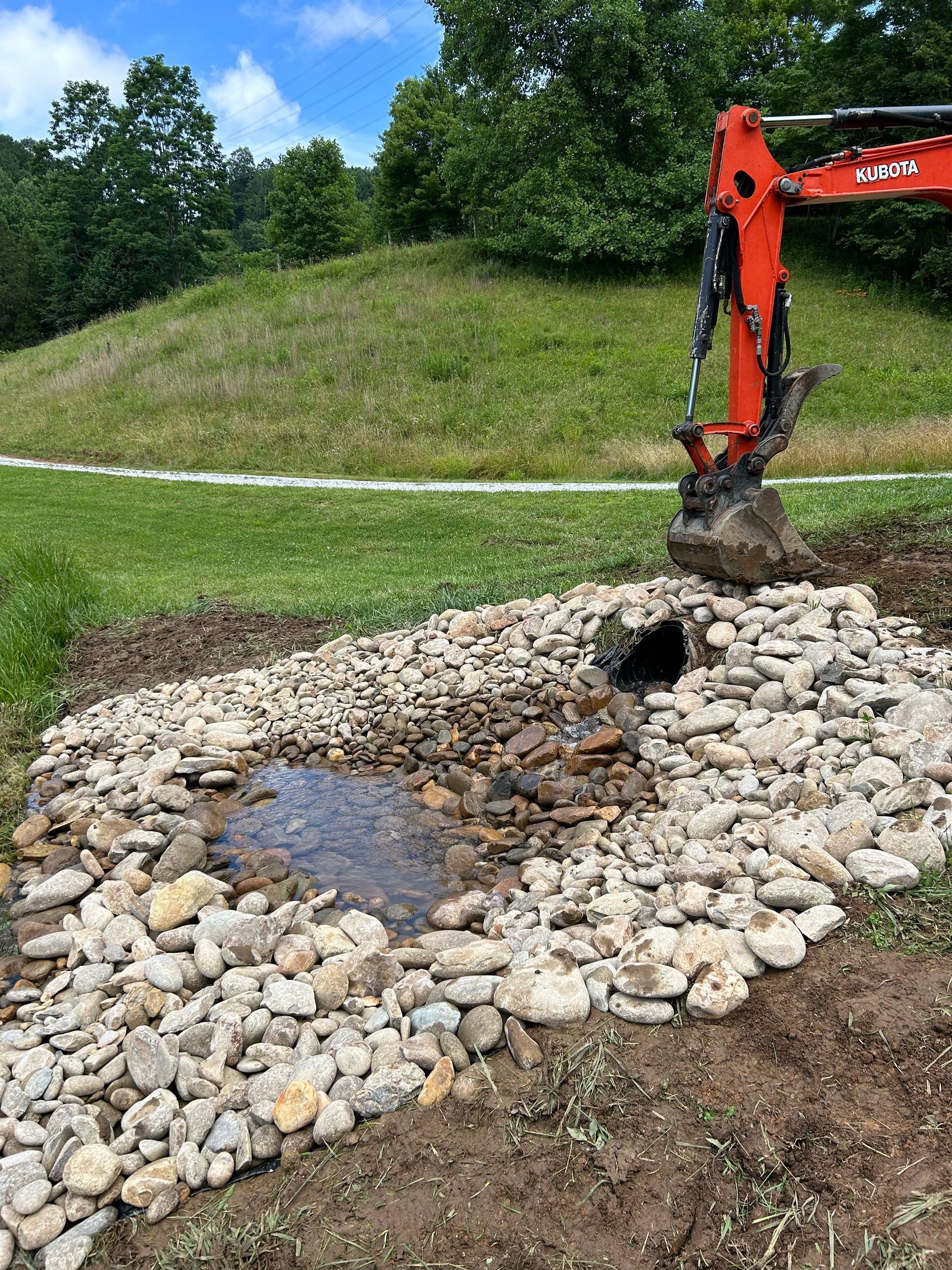 Orange excavator digging in a rocky drainage area beside a grassy hill and trees.