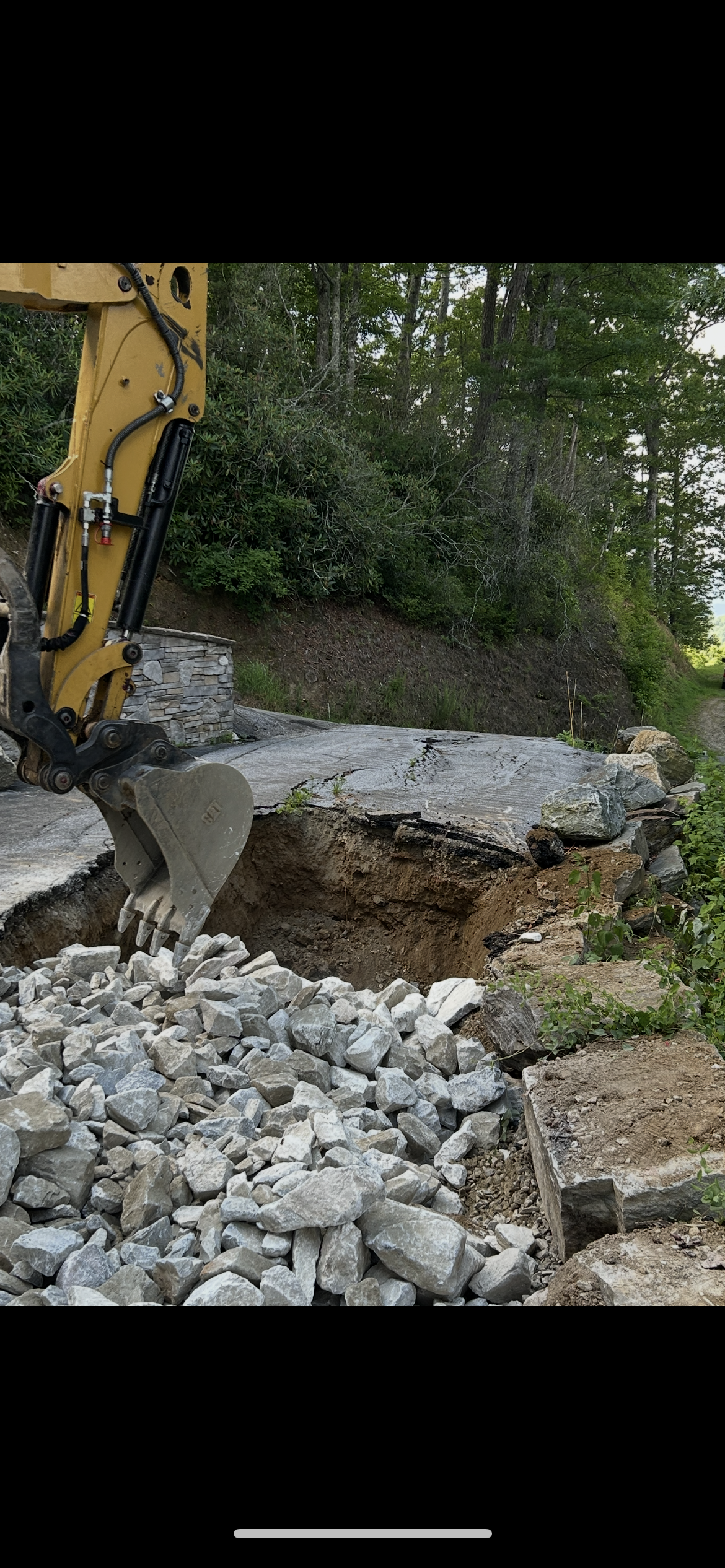 Yellow excavator arm demolishing a large concrete slab amid rubble and trees outdoors