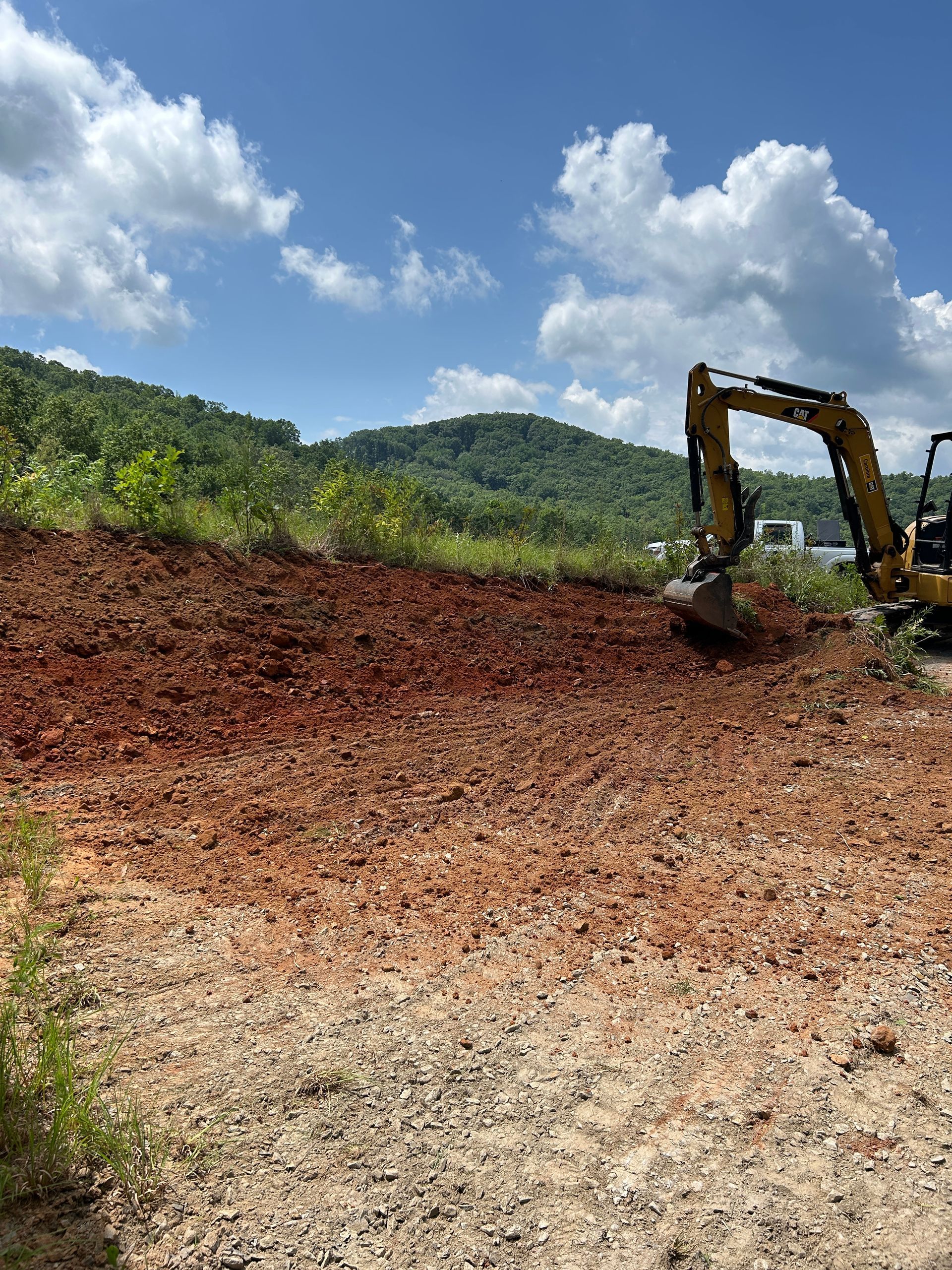 Excavator clearing red soil on a hillside under a blue sky with clouds.