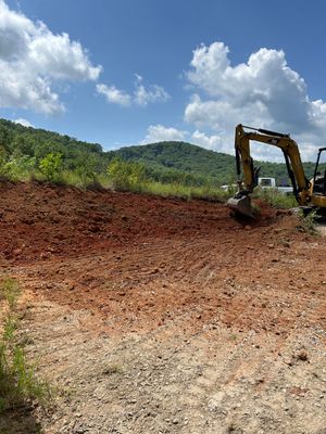 Excavator grading red dirt on a hillside under a blue sky with clouds