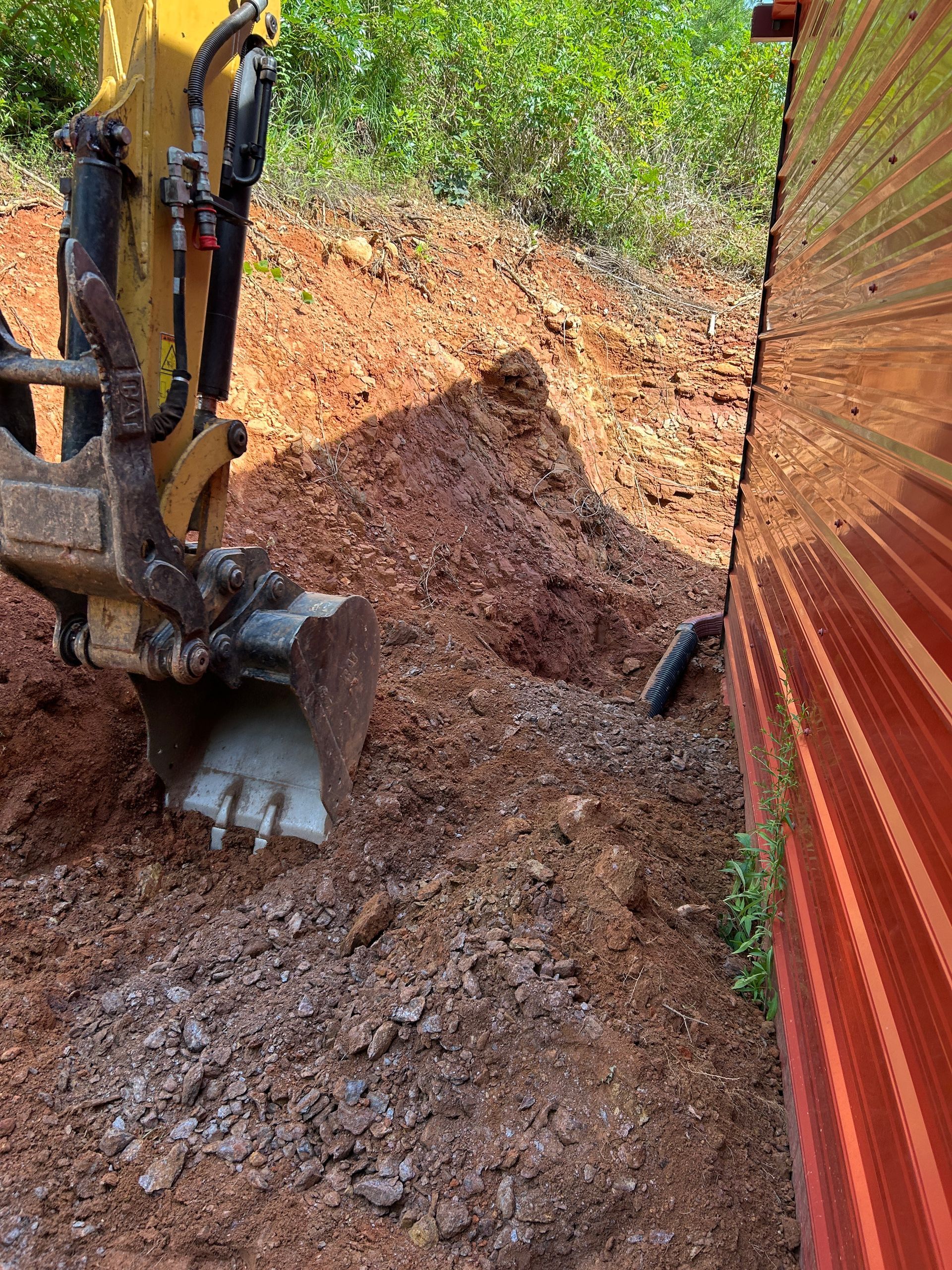 Excavator bucket digging a dirt trench beside a red corrugated wall in a wooded area