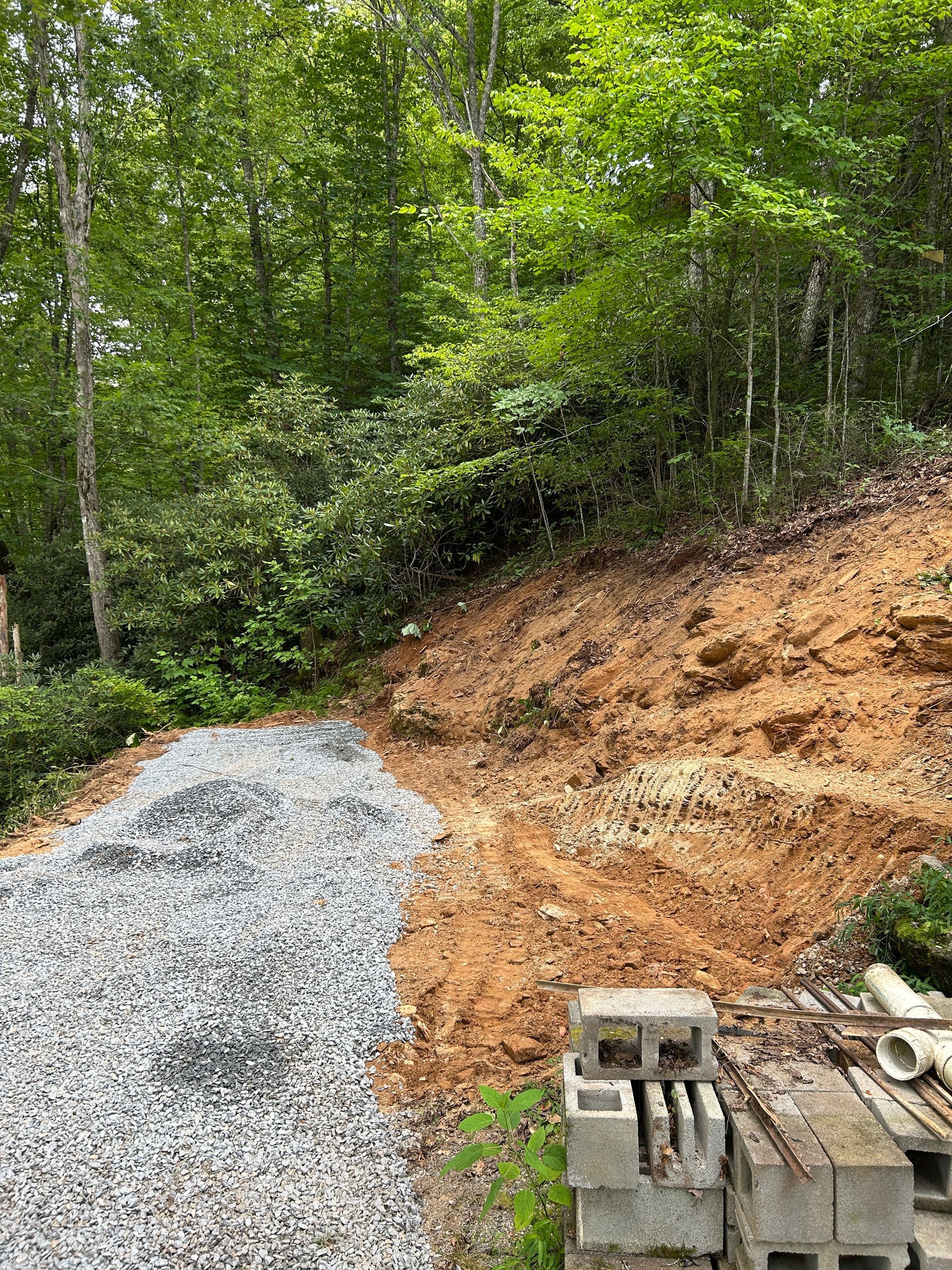 Gravel driveway beside a wooded hillside with exposed orange soil and stacked cinder blocks in the foreground
