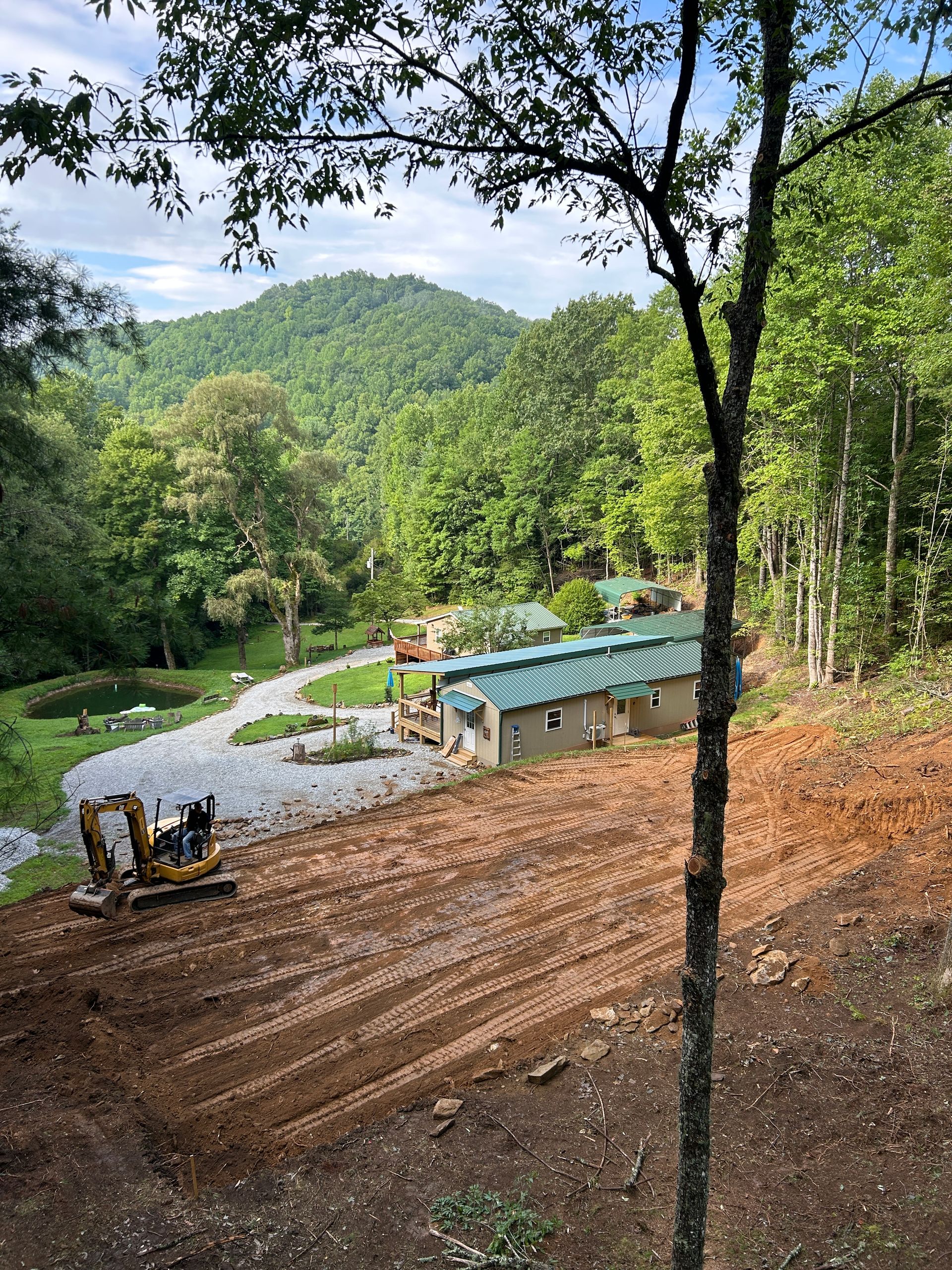 Forest hillside with a winding gravel road, small blue-roofed shelter, and machinery near trees and hills.