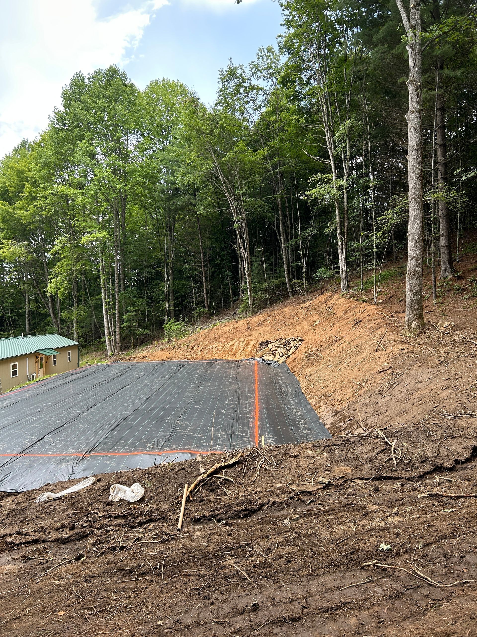 Gravel driveway under construction beside a wooded hillside with soil slope and a small building in the background