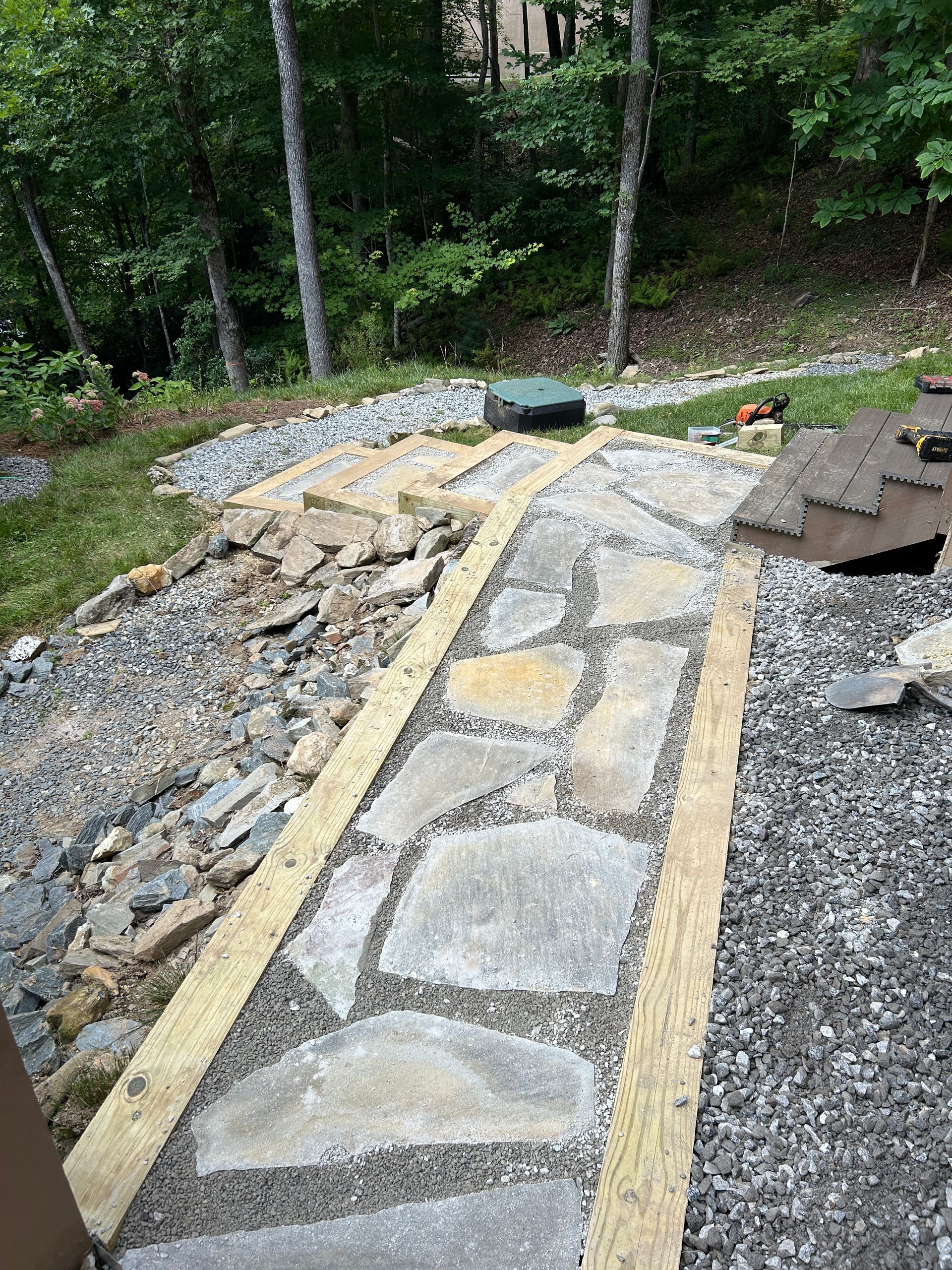 Stone walkway under construction in a wooded yard, with gravel, edging boards, and a stepped path layout