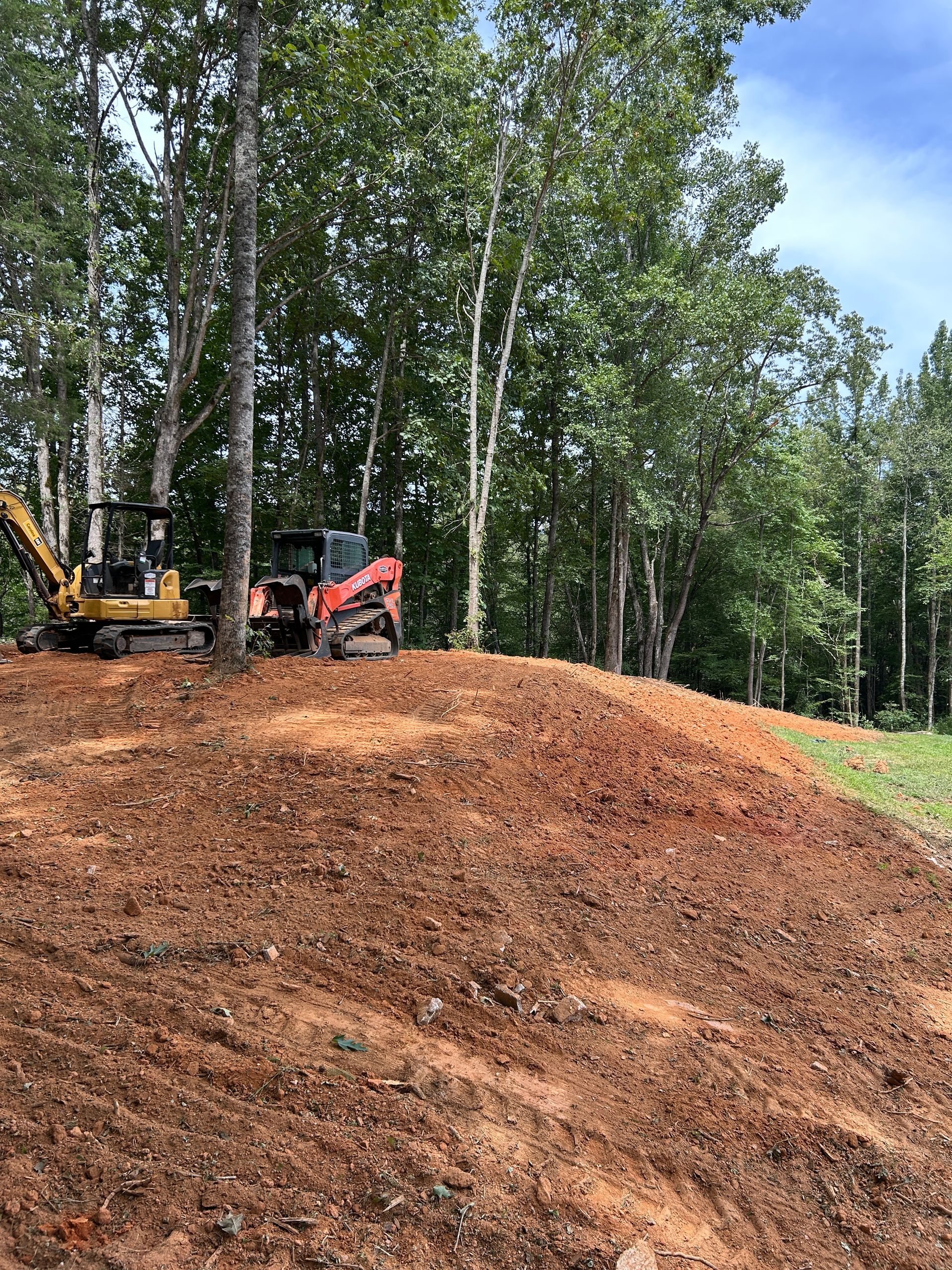 Excavator and red skid steer on a dirt mound at a wooded construction site under a clear sky