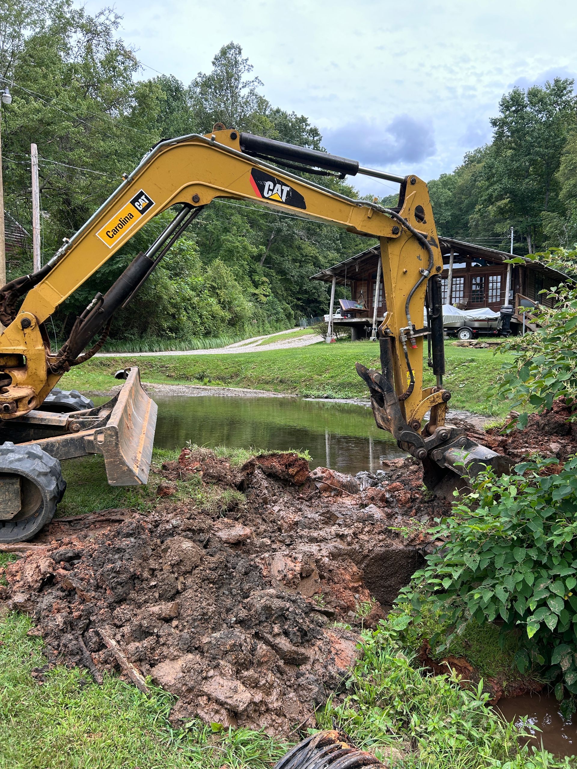 Yellow excavator digging muddy earth beside a pond in a wooded yard