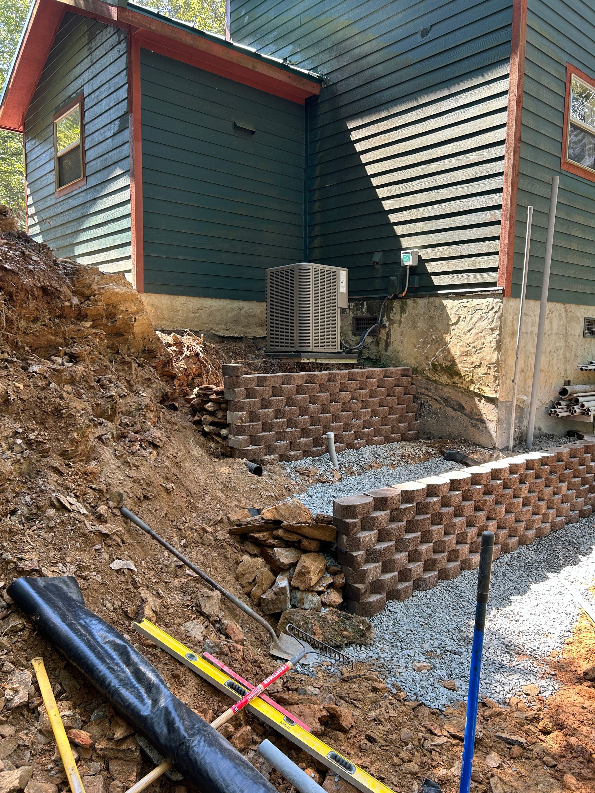 Side yard beside a house with a retaining wall, exposed soil, gravel, and construction tools on the ground