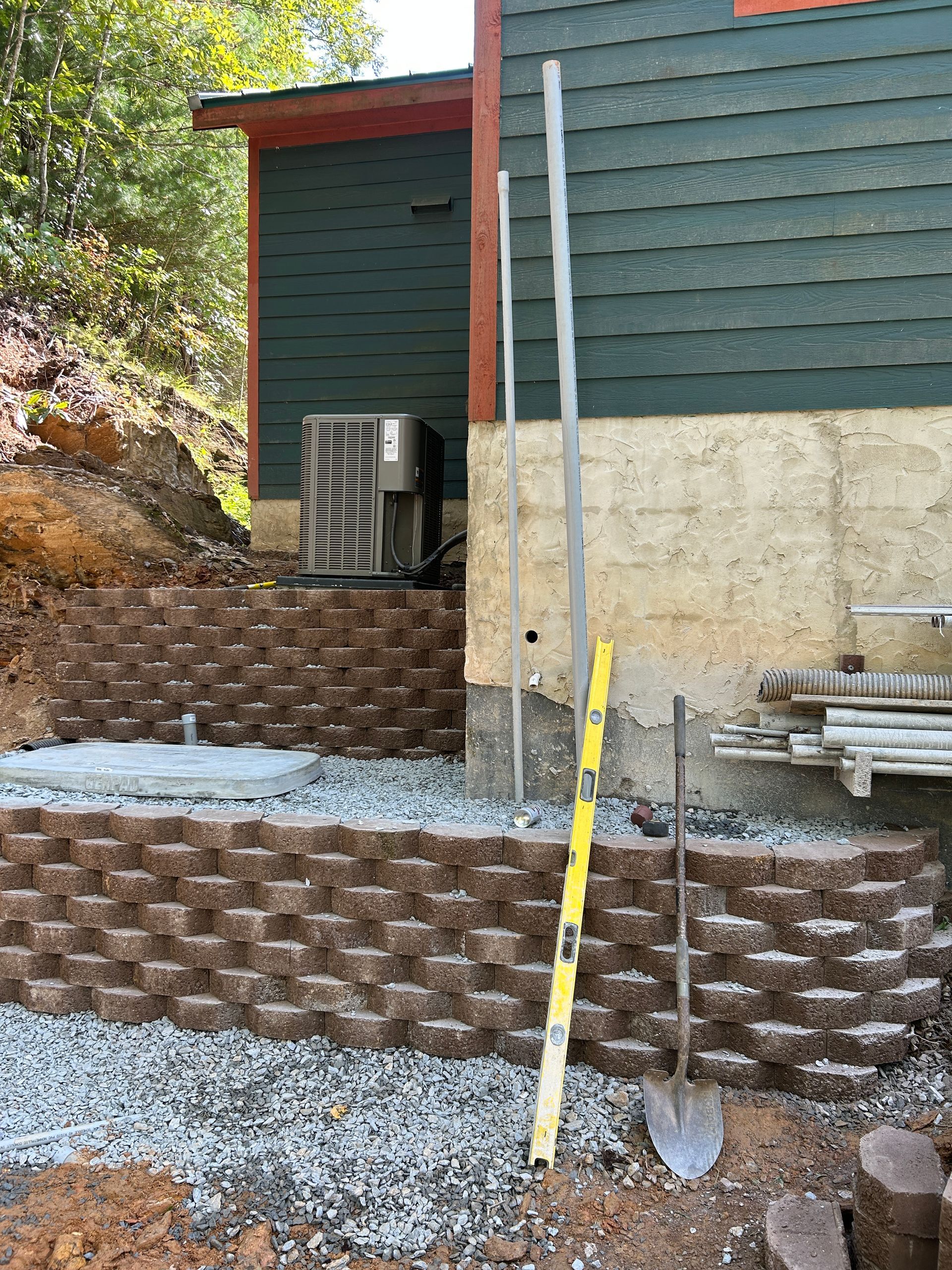 Backyard retaining wall under construction beside a house, with shovel and level leaning against it.