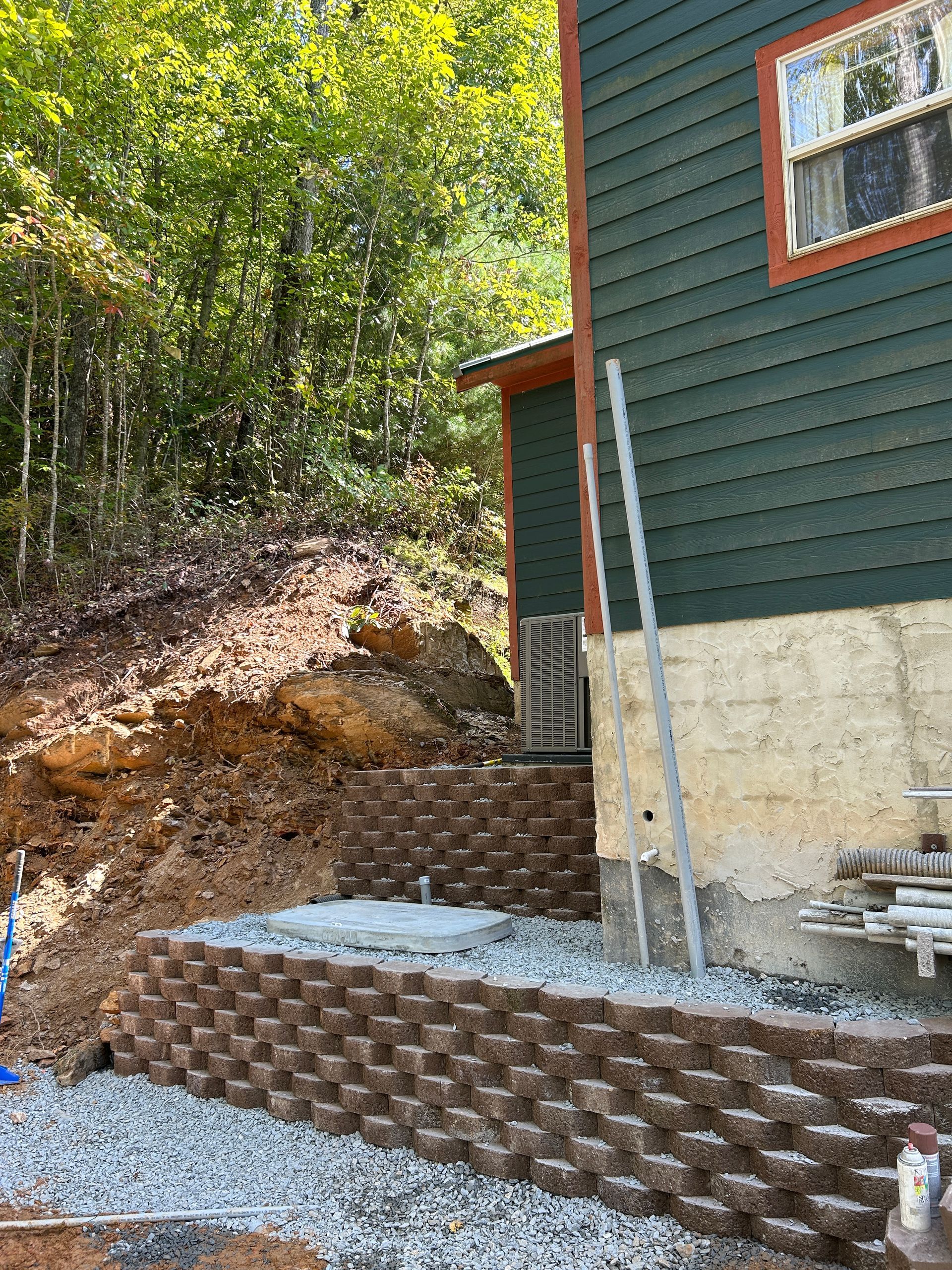 Exterior of a house with green siding, new stone retaining wall, and a dirt slope beside wooded area