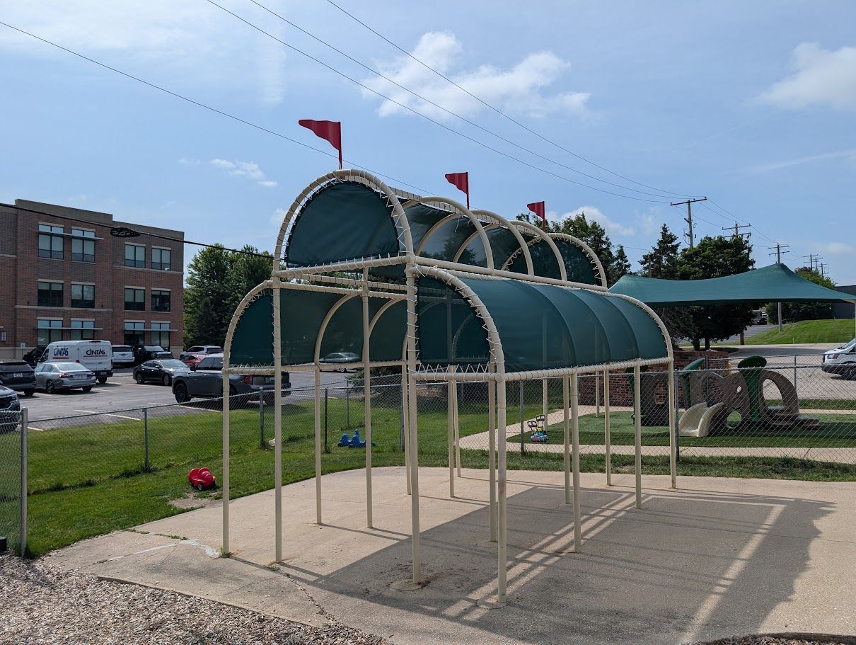 Covered playground structure with green canopy and red flags on a sandy lot near buildings
