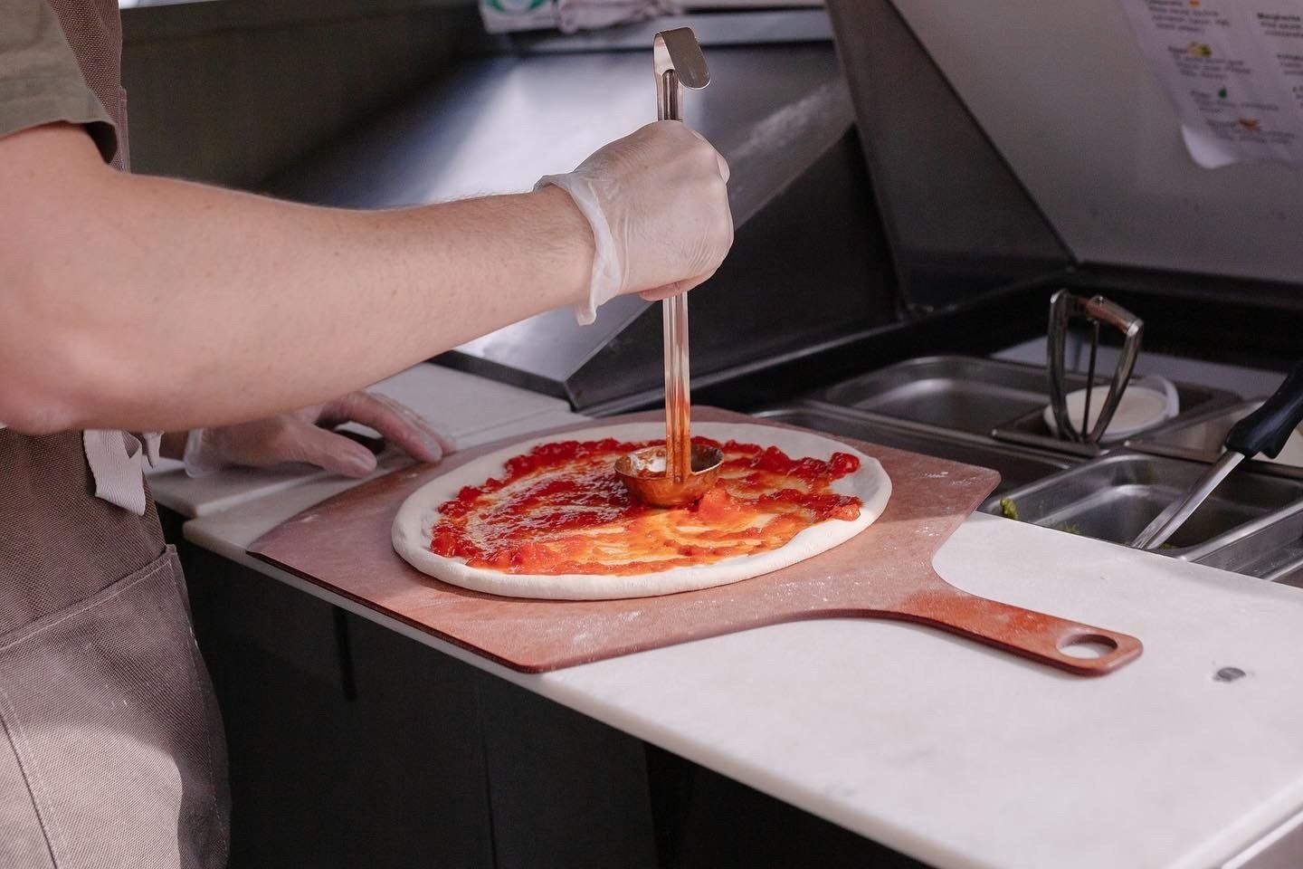 A person is spreading sauce on a pizza with a spatula at Beyond Basil Mobile Pizzeria.