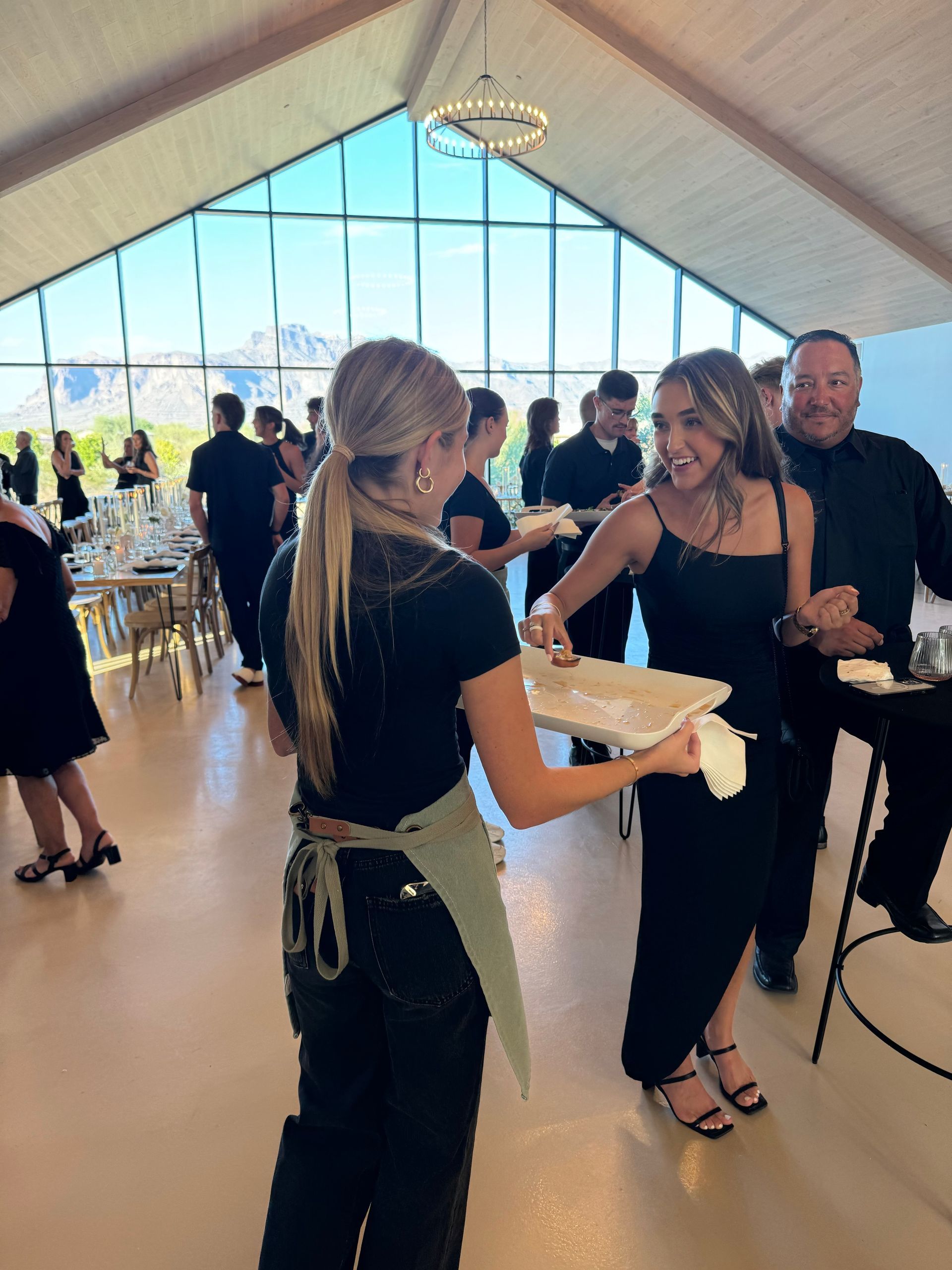 Server handing a wooden board with food to a person in a black dress at a party. Bright room with tables.