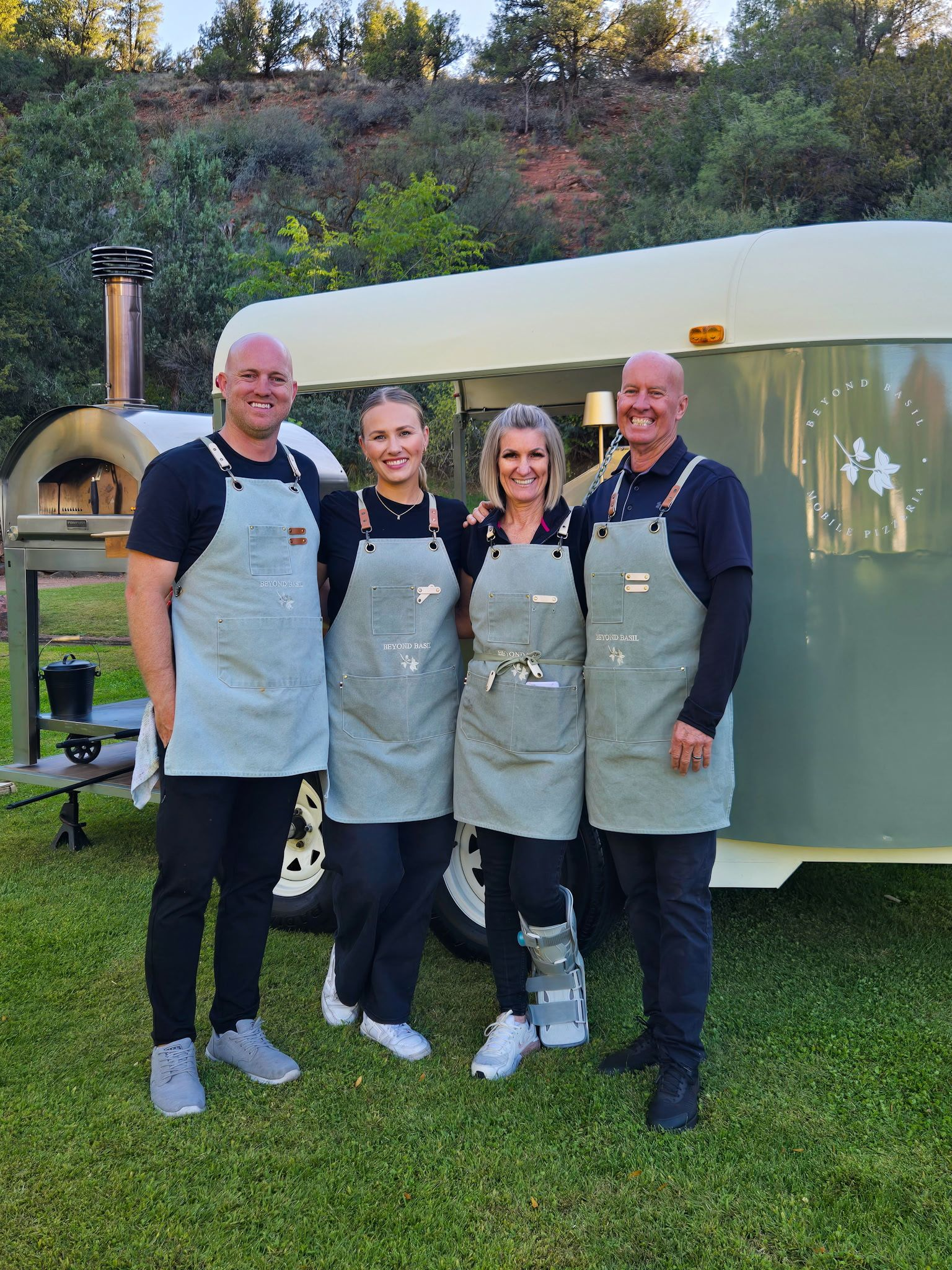 Four people in aprons stand in front of a pizza oven and trailer, posing on grass.