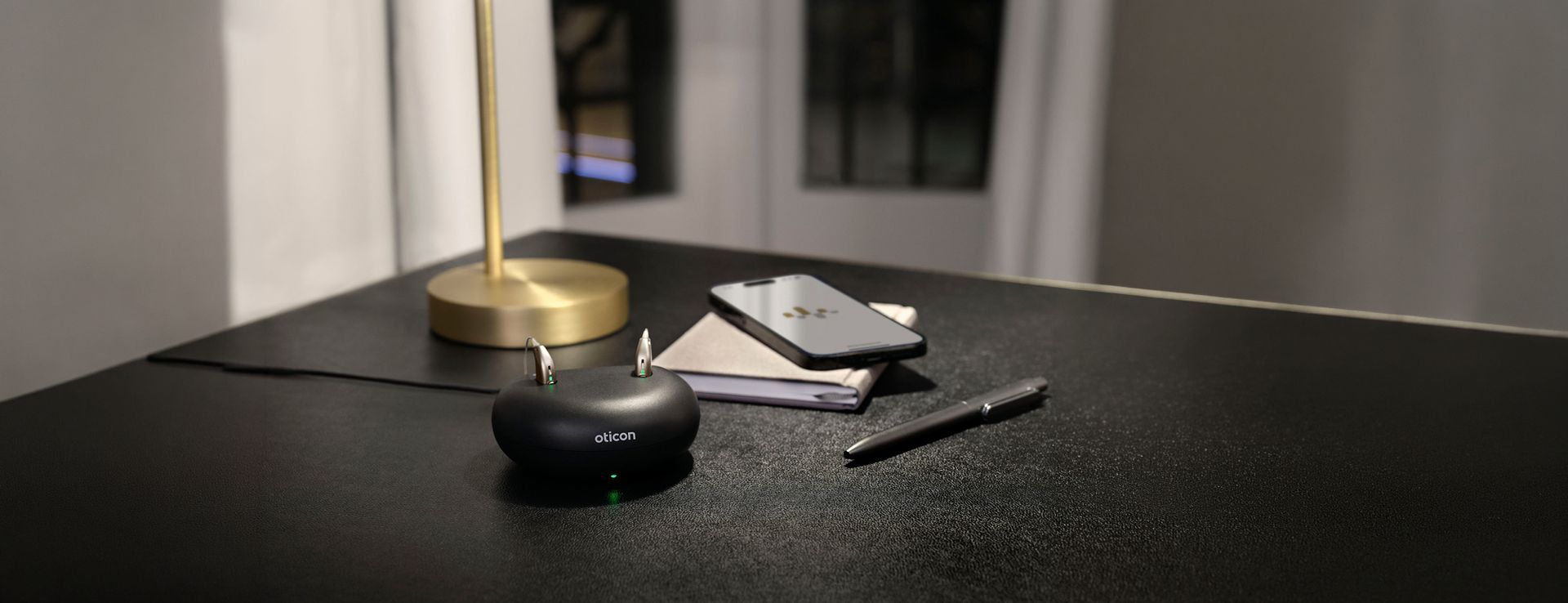 Oticon hearing aids charging in a wireless charger on a desk beside a smartphone