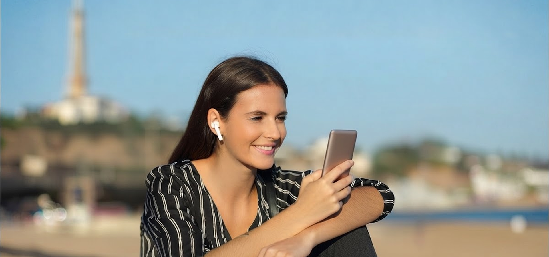 Woman wearing a single wireless earbud smiling at her smartphone outdoors on a sunny beach