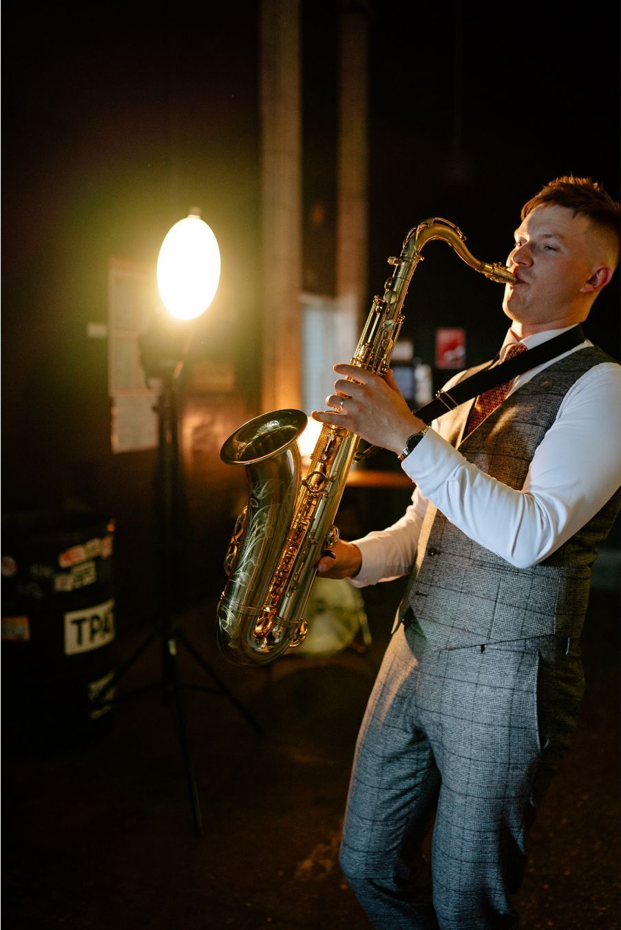 Man playing saxophone on a dimly lit stage, wearing a white shirt and gray vest, with a bright light behind him