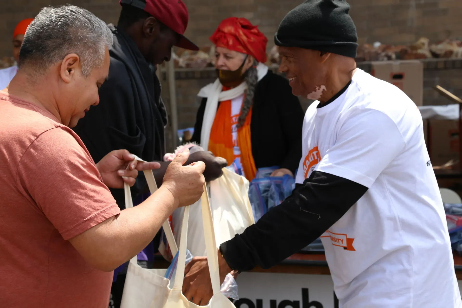 People receiving food bags at an outdoor event. Man in white shirt hands a bag to another person.