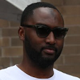 A man wearing sunglasses and a white shirt is standing in front of a brick wall.
