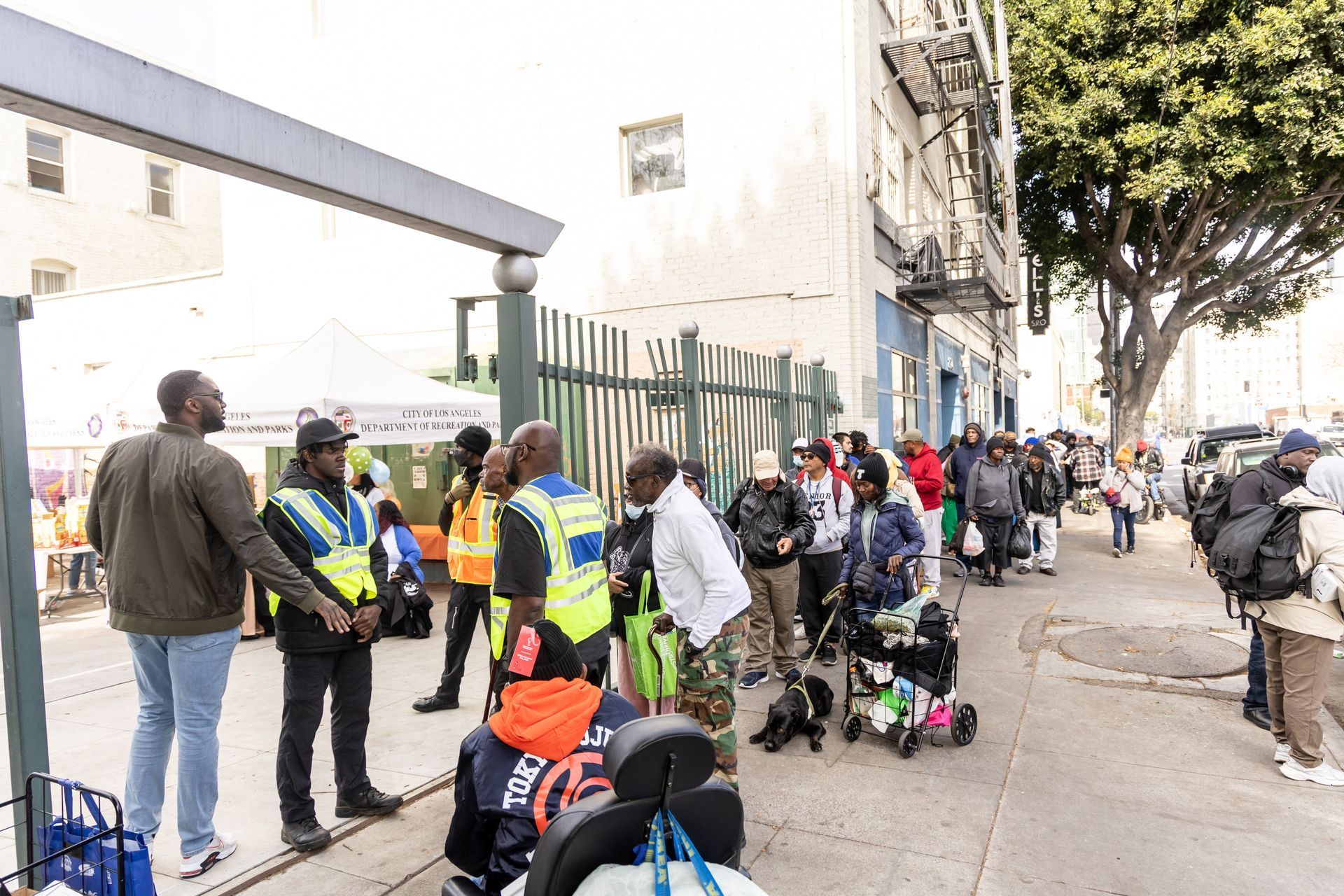 People line up outside a building, some with bags and a dog. Security guards stand nearby.