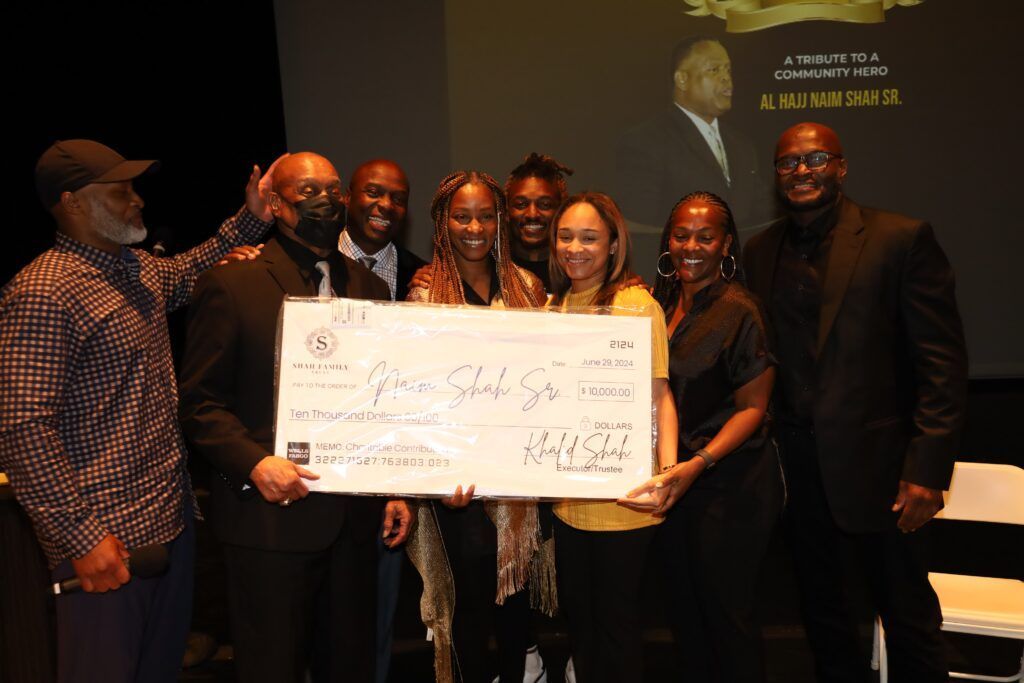 Group holding large check, celebrating; dark attire, onstage with screen.