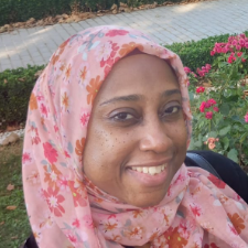 Woman in a pink floral hijab smiles outdoors near greenery.