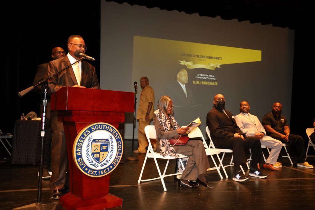 Man speaking at a podium, with a presentation screen and seated audience on stage.