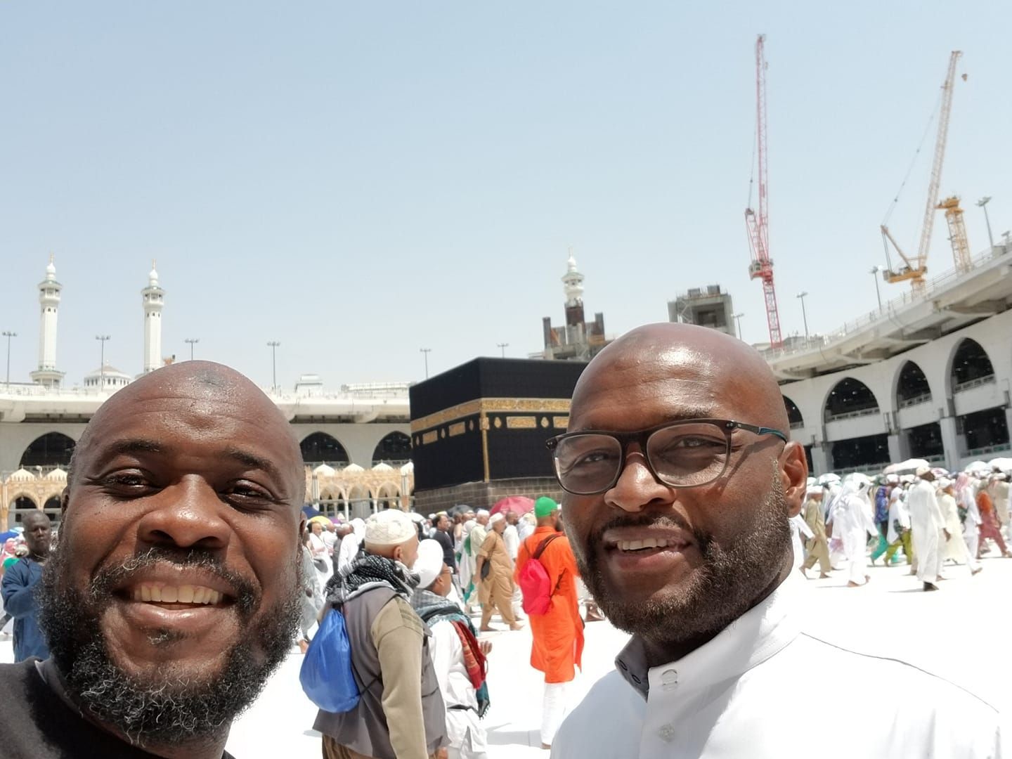 Two men smiling in front of the Kaaba at the Grand Mosque in Mecca, with many people in the background.