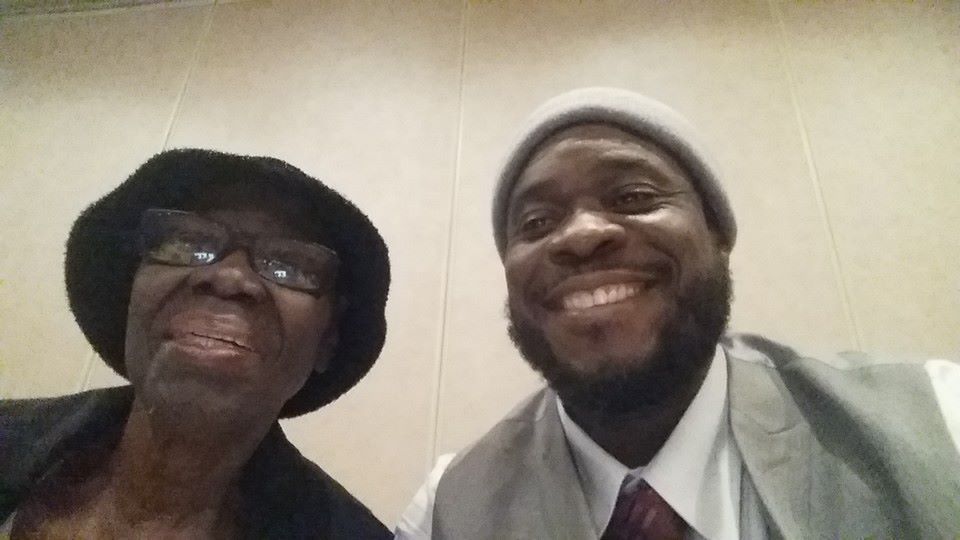 Two people smiling at the camera. One wears a hat and glasses, the other a beanie and vest.